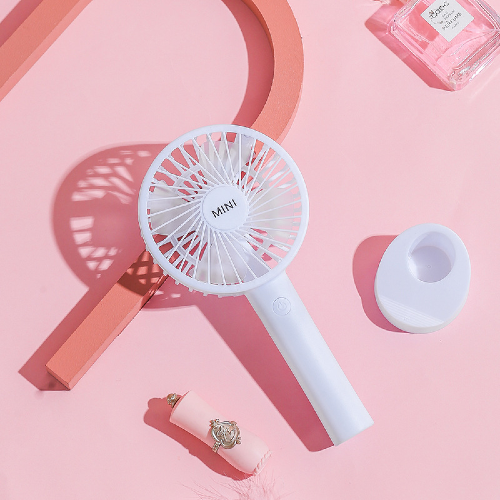Ein weißer Mini-Ventilator, pinker Lippenstift und eine Parfümflasche auf pinkem Hintergrund.