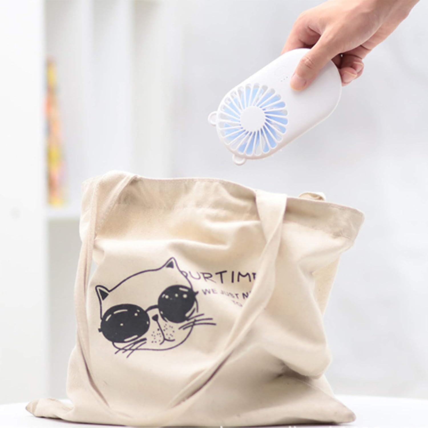 Eine Hand hält einen kleinen weißen Ventilator über einer cremefarbenen Stofftasche mit Katzenaufdruck.