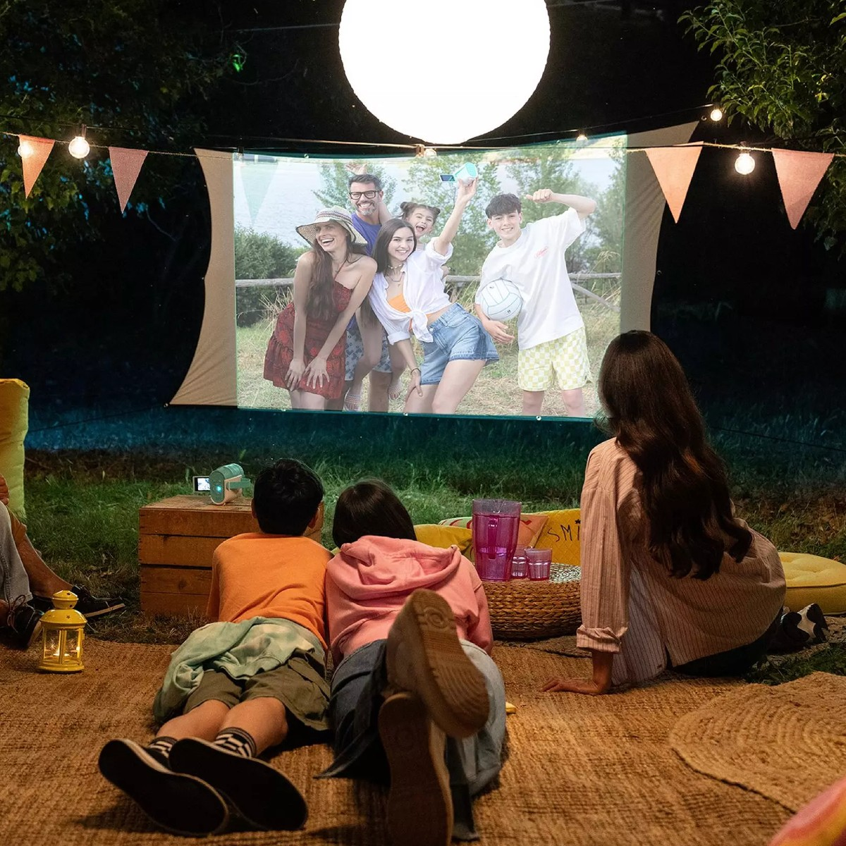 Gente viendo una película al aire libre en una pantalla, iluminada por luces y un orbe grande.