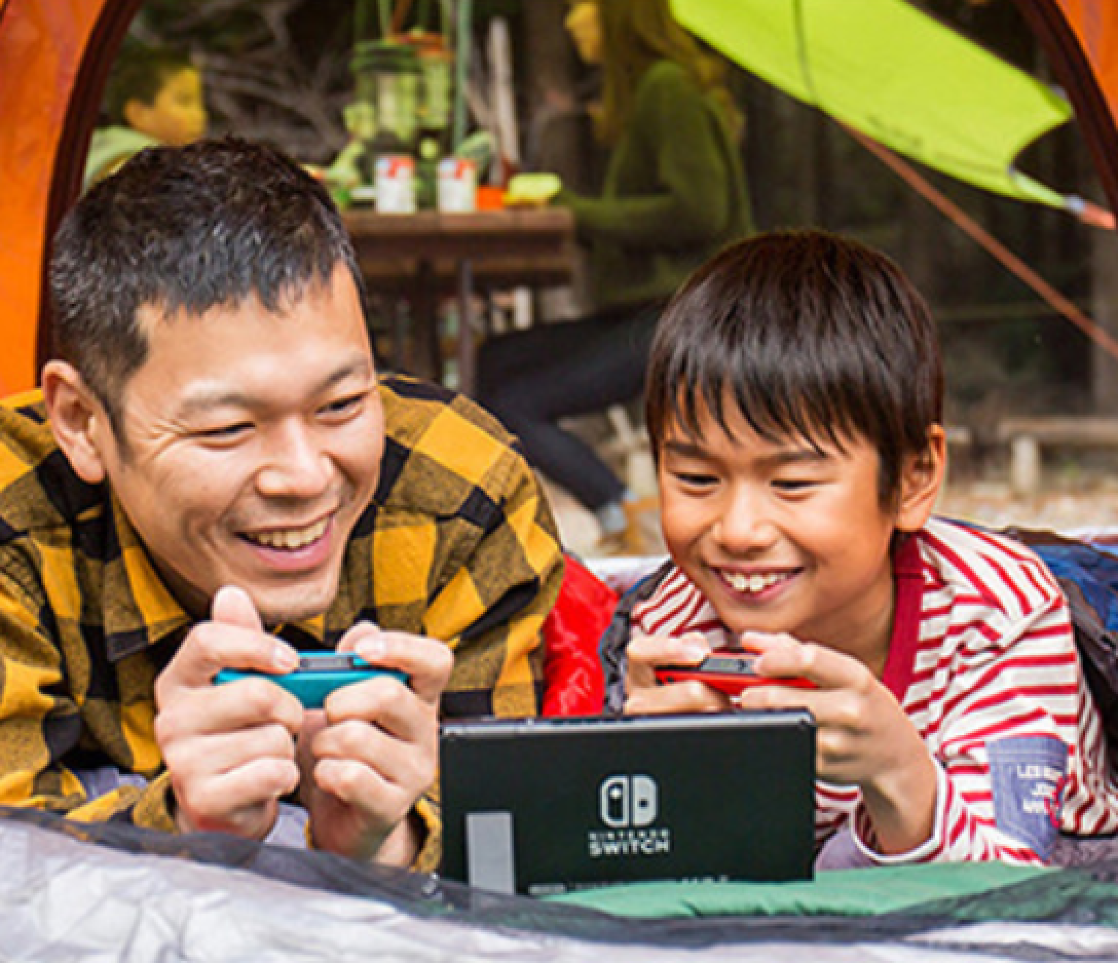 Vater und Sohn spielen Nintendo Switch in einem Zelt mit Blick auf andere Camper.