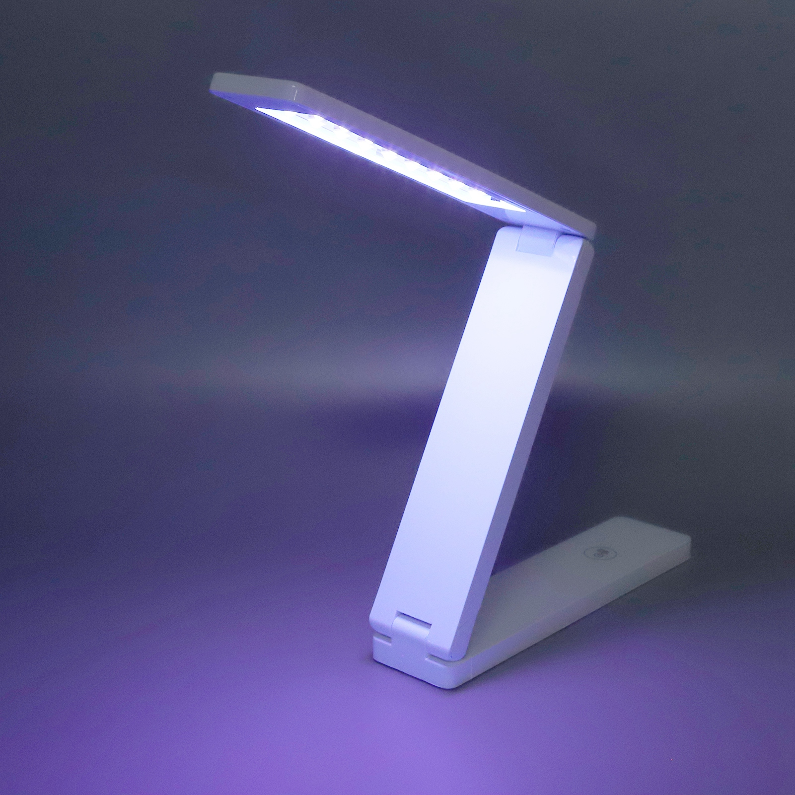 Eine weiße, faltbare LED-Schreibtischlampe mit violettem Licht leuchtet auf einem grauen Hintergrund.