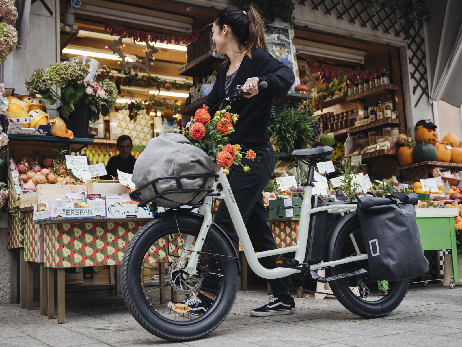 Eine Frau schiebt ein Fahrrad mit Blumen und Beutel im Korb vor einem Gemüsestand.