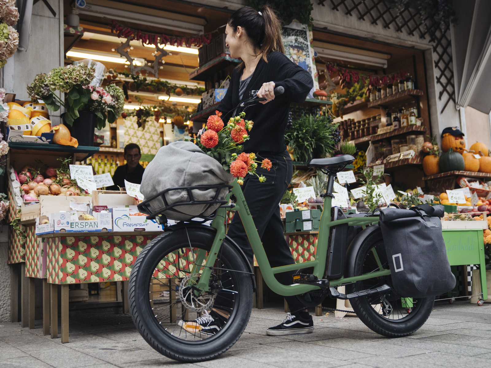 Eine Frau schiebt ein grünes Fahrrad mit Blumenkorb vor einem Marktstand mit Obst und Gemüse.