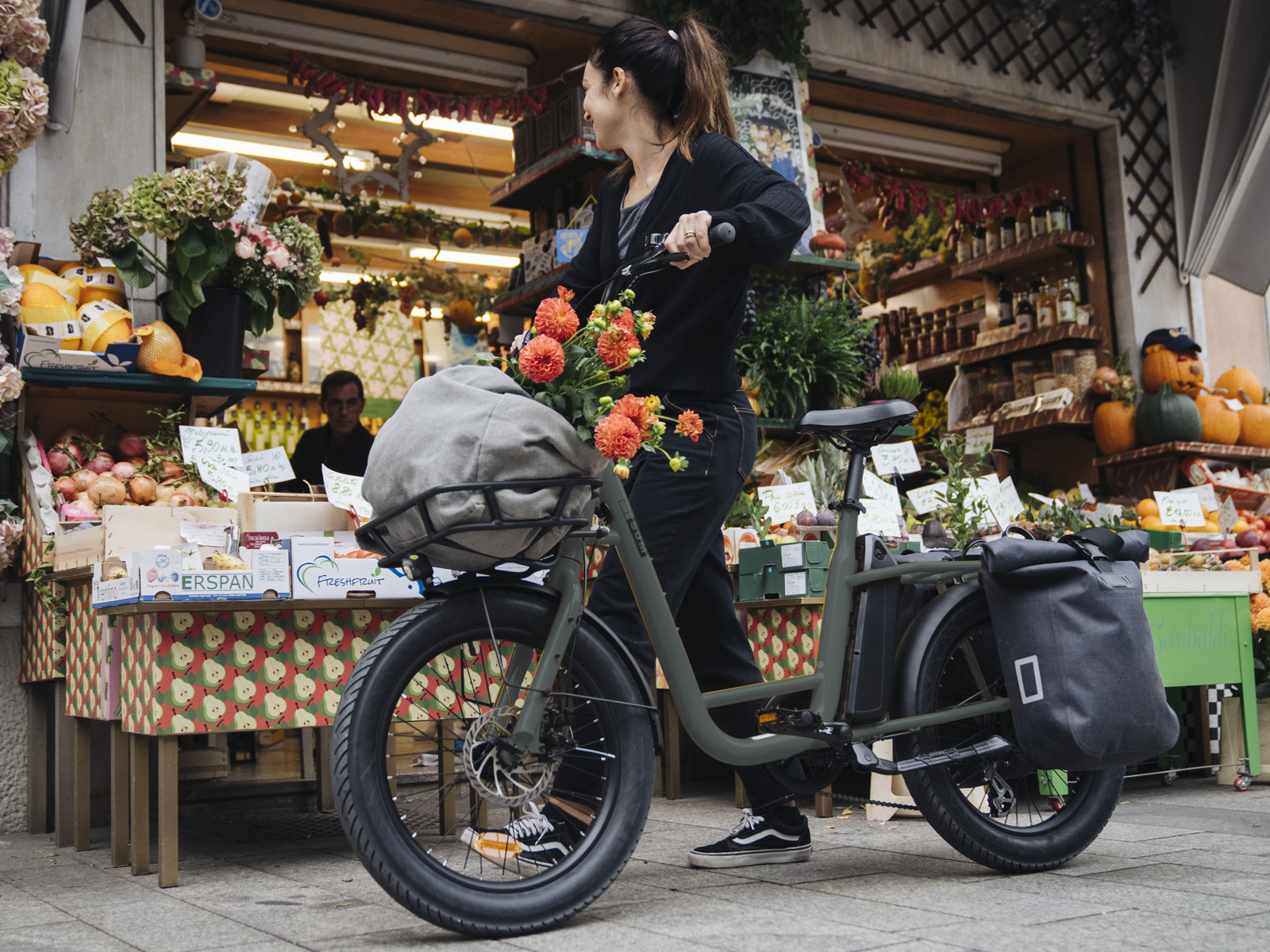 Eine Frau schiebt ein Fahrrad mit Blumen im Korb vor einem Obststand.