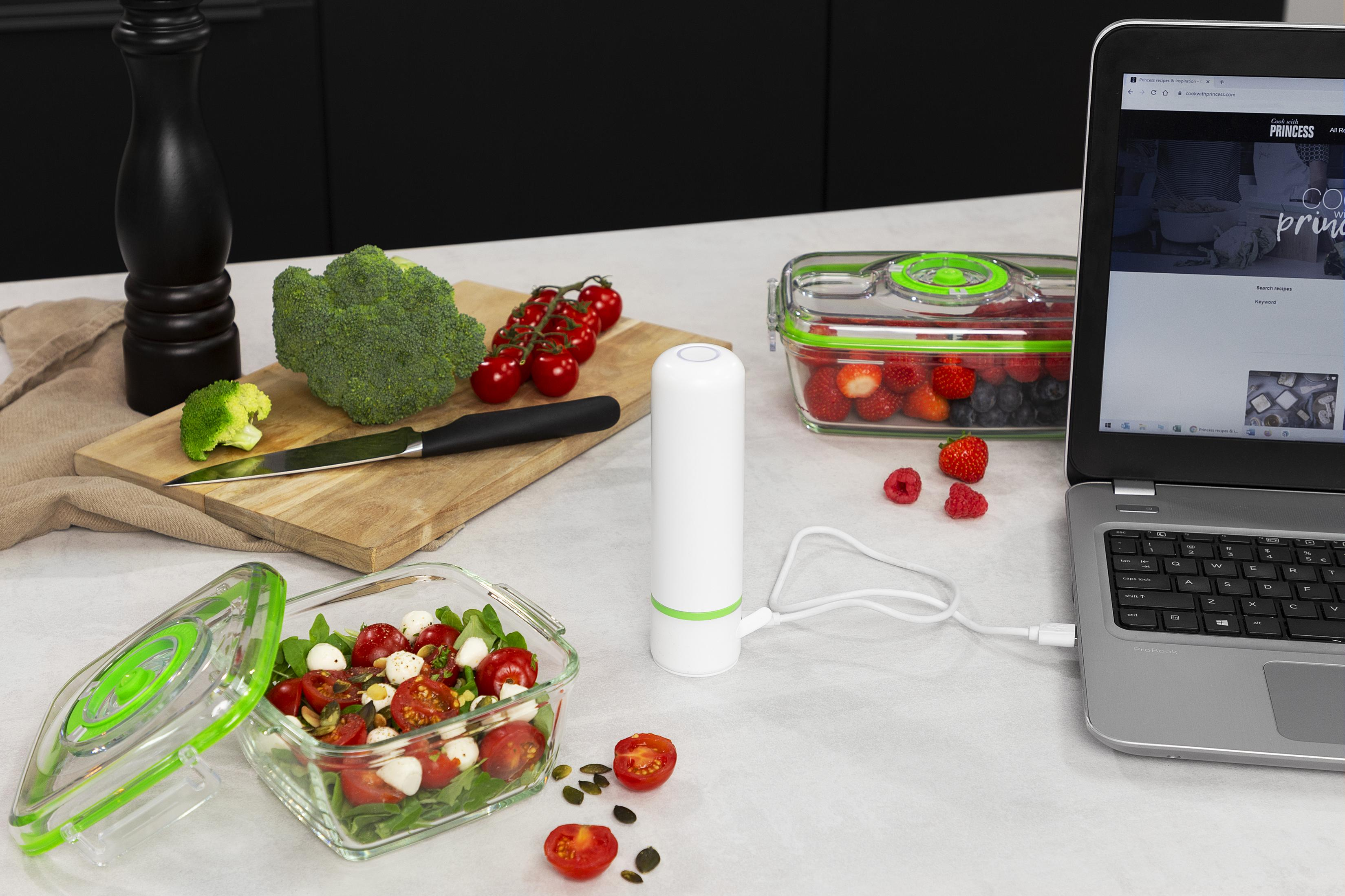 Ein Vakuumiergerät lädt neben Beeren und einem Salat, auf einem Laptop mit Rezeptseite.