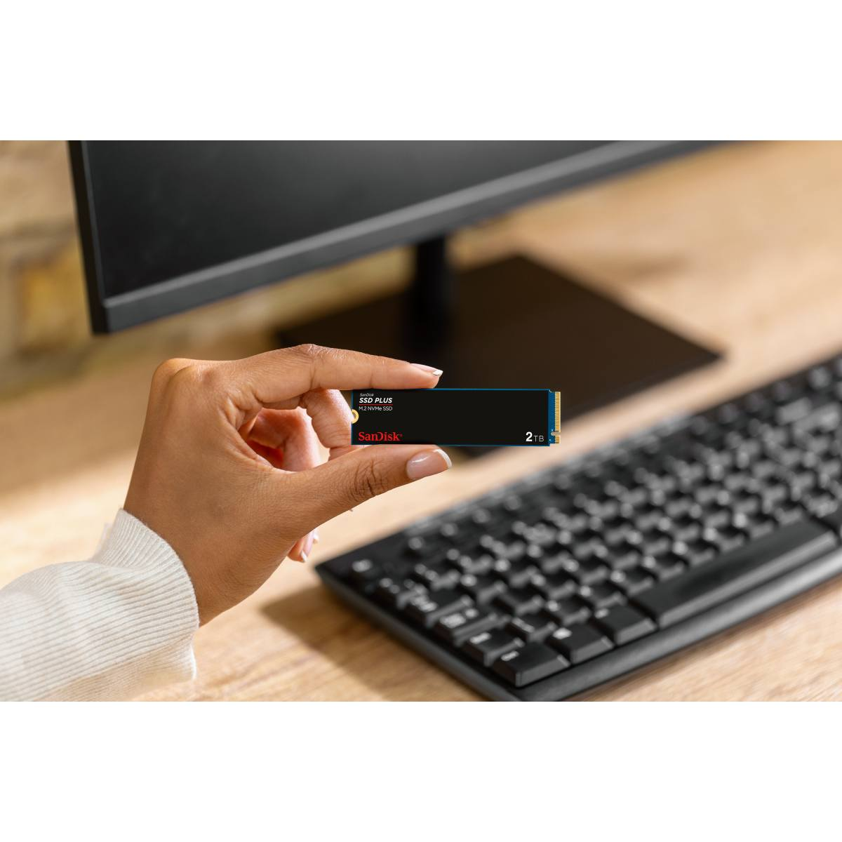 Ręka trzyma czarny dysk SanDisk SSD Plus 2TB M.2 NVMe przed monitorem.