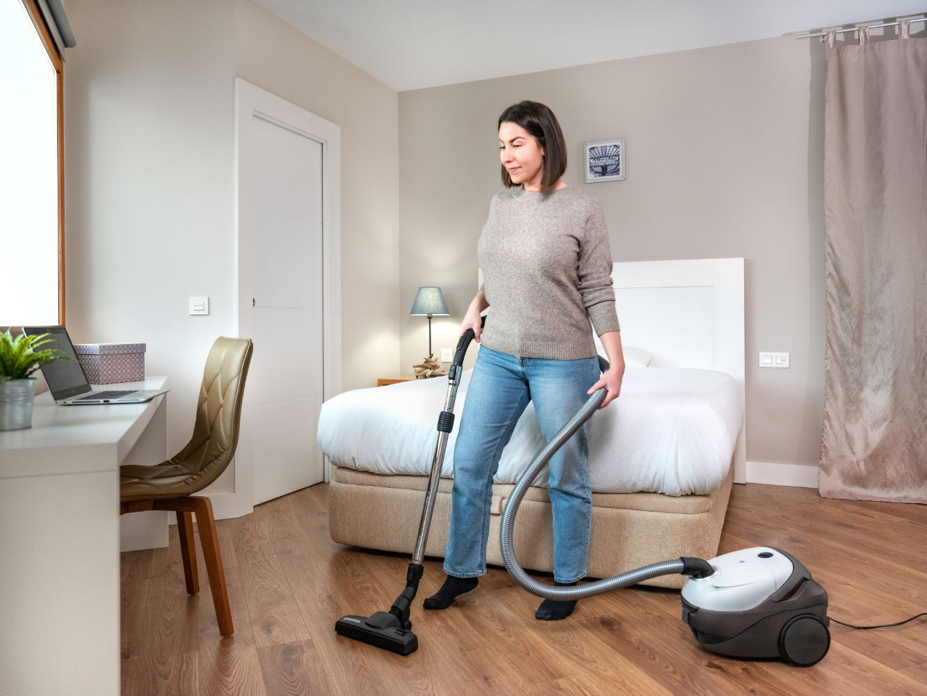 Une femme passe l'aspirateur sur un parquet dans une chambre avec un lit et un bureau.