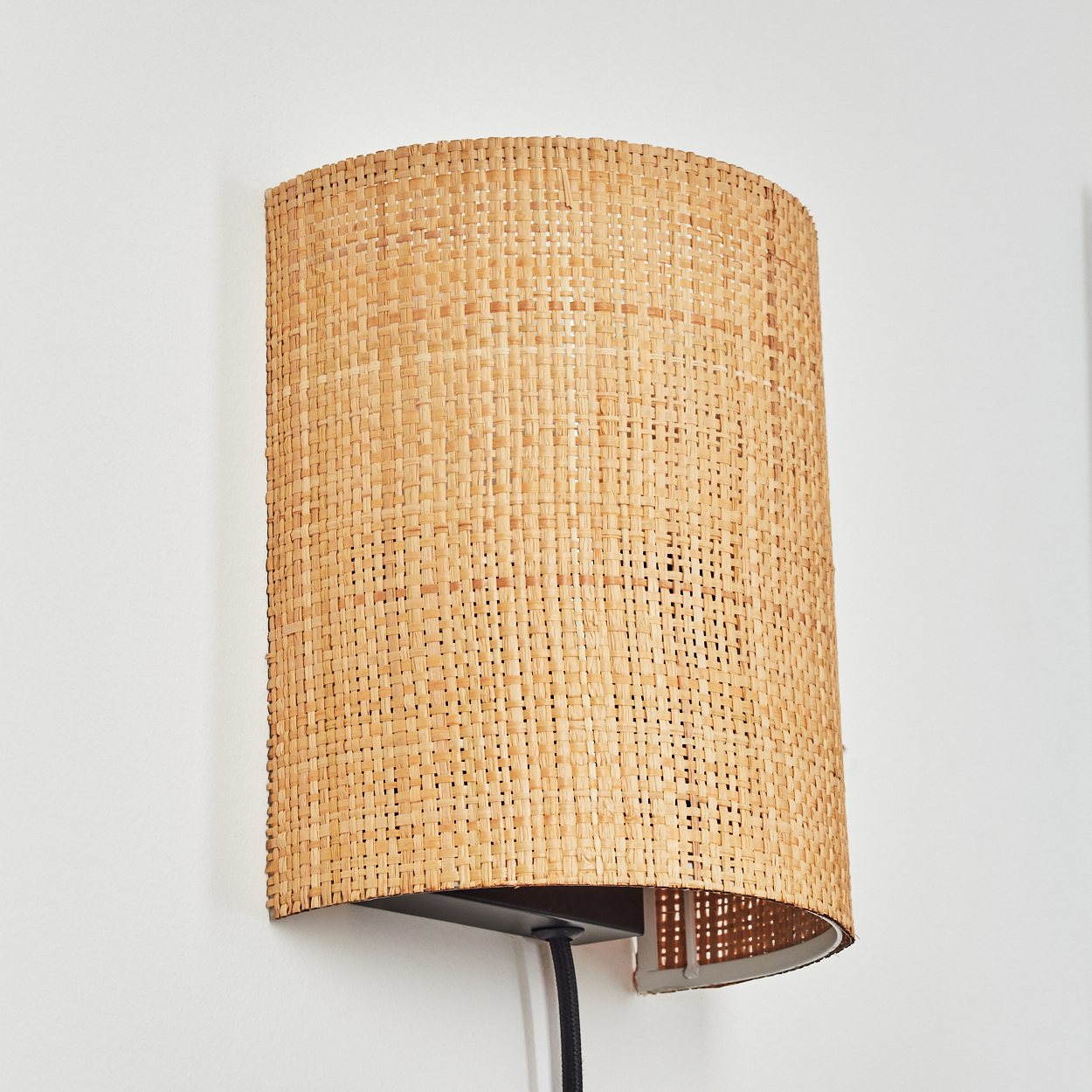 Eine Wandleuchte mit einem Korbrohrschirm auf weißer Wand, schwarzes Kabel.