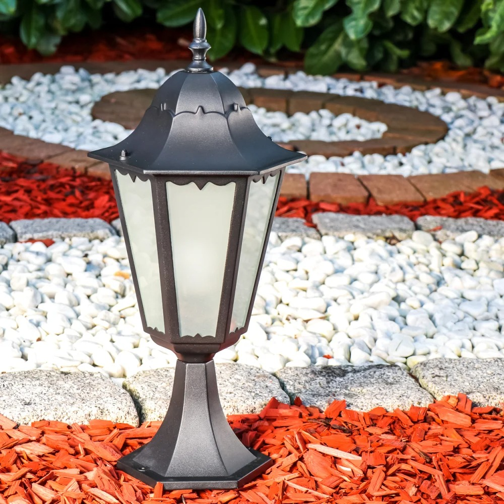 Eine schwarze Gartenlampe steht in roter Rindenmulch, weißen Steinen und Grün.
