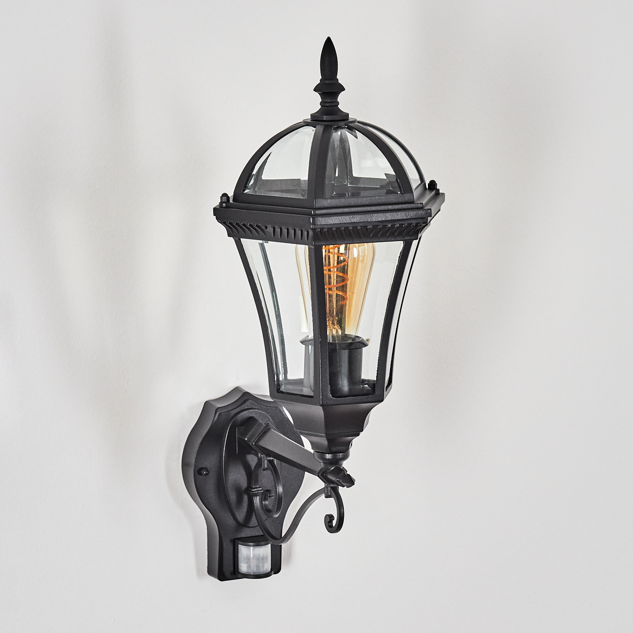 Schwarze Außenlaterne mit Glasscheiben und Edison-Birne, an einer weißen Wand montiert.
