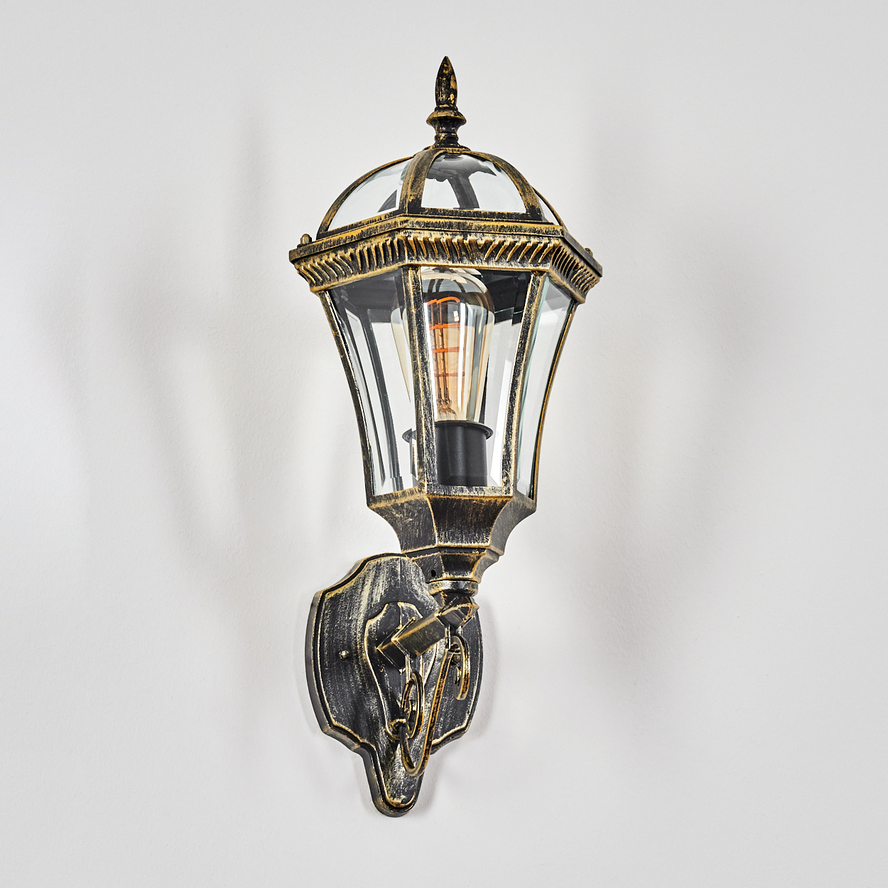 Eine Wandleuchte im Laternenstil mit gold- und schwarzem Finish, Glas und Glühbirne.