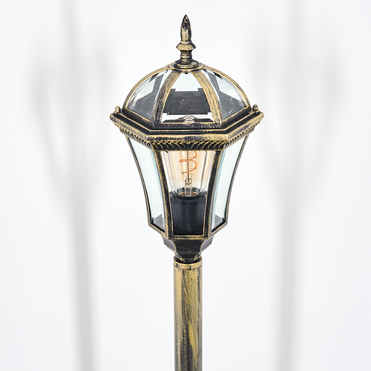 Eine goldene Lampe mit Glaseinsätzen und sichtbarer Glühbirne vor weißem Hintergrund.