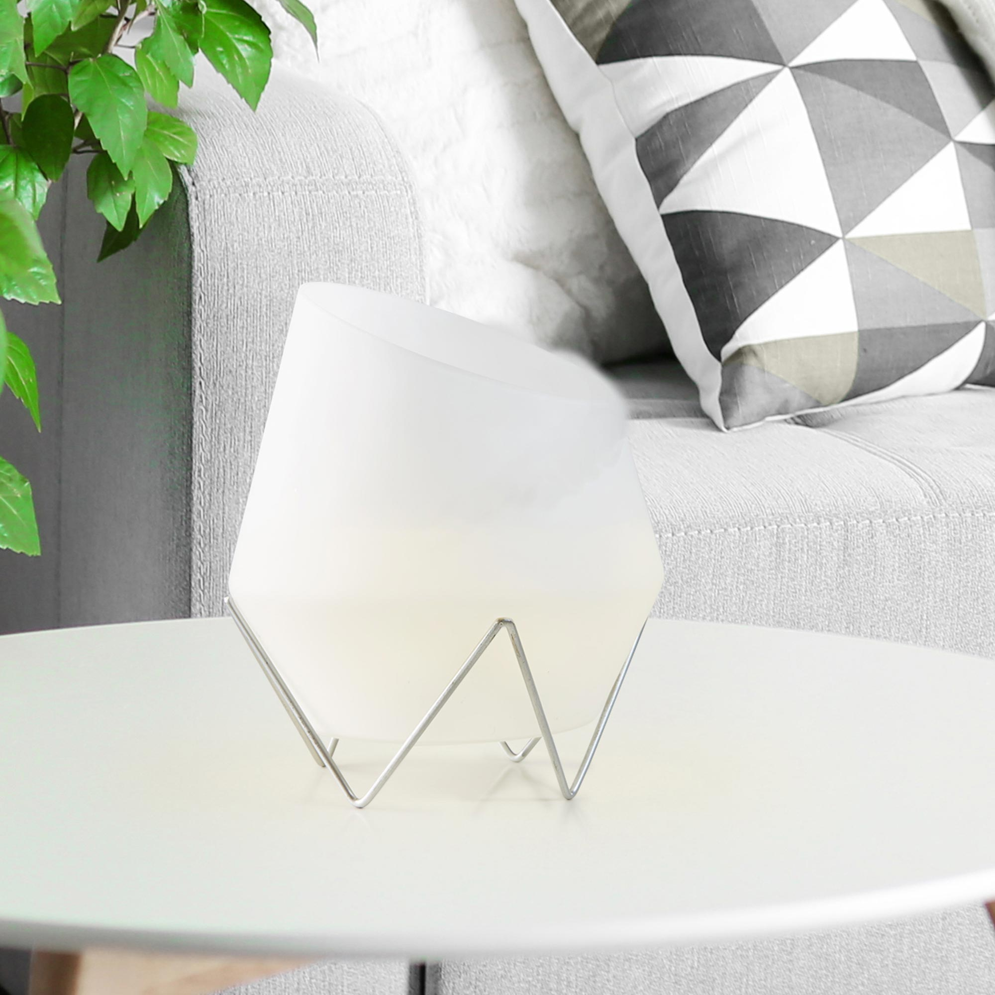 Une lampe blanche sur pied métallique repose sur une table devant un canapé avec coussins.