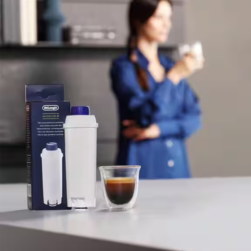 Eine Frau in einem blauen Hemd hält ein Glas, neben einer Kaffeemaschine und einem Espresso.