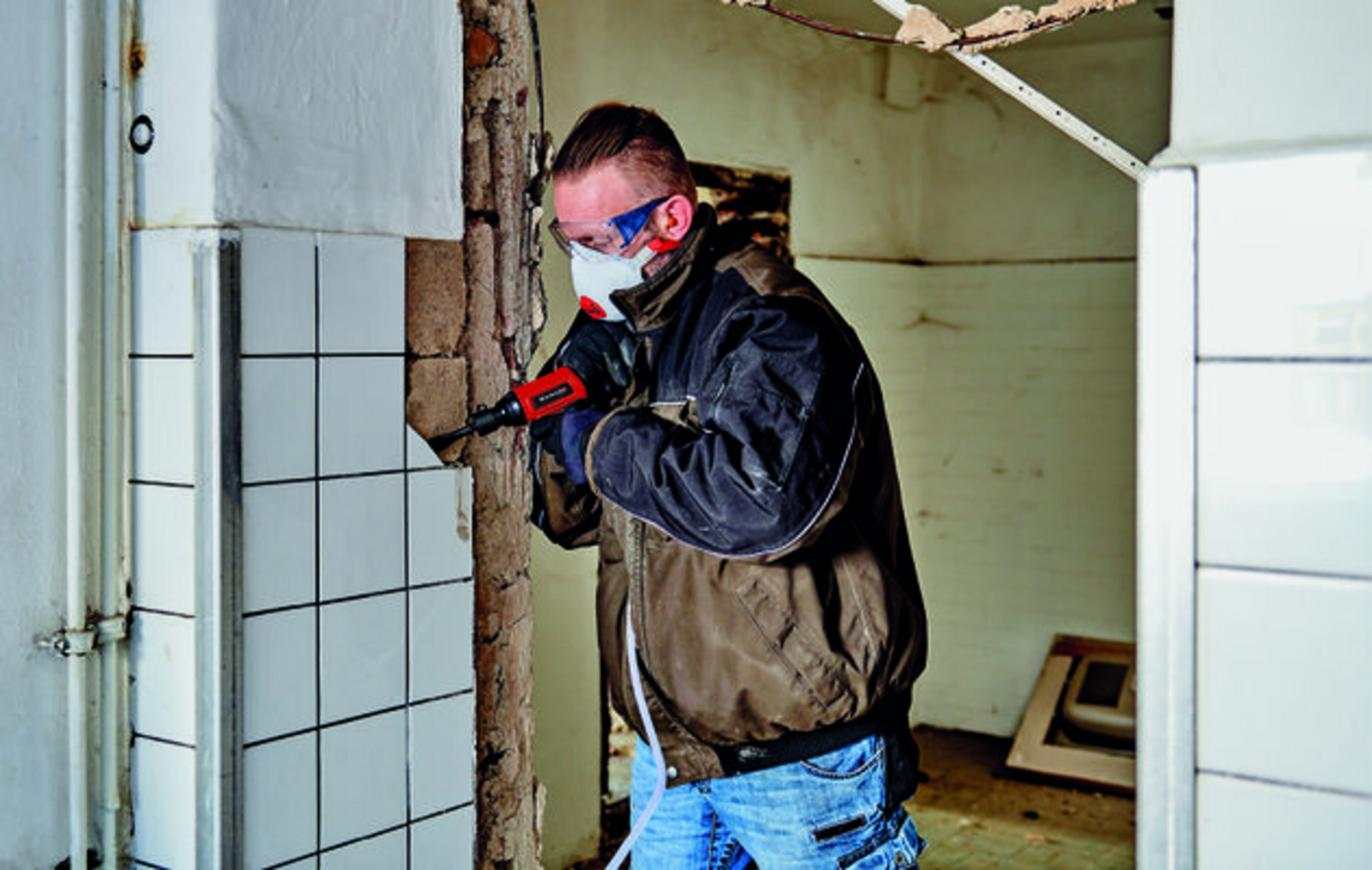 Uomo con maschera e occhiali rompe muro con attrezzo.