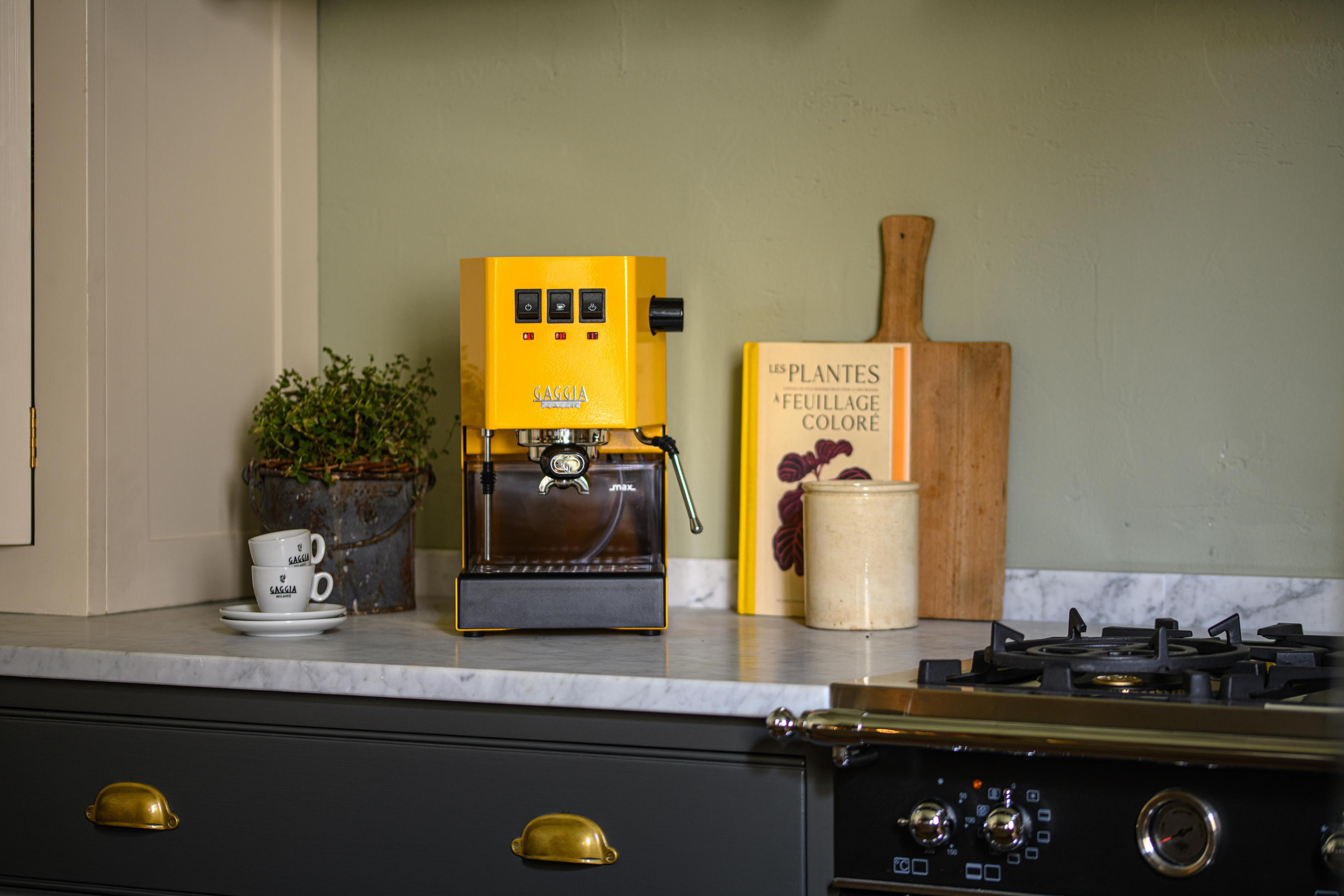Een gele espressomachine staat op een marmeren aanrecht naast een potplant en boeken.