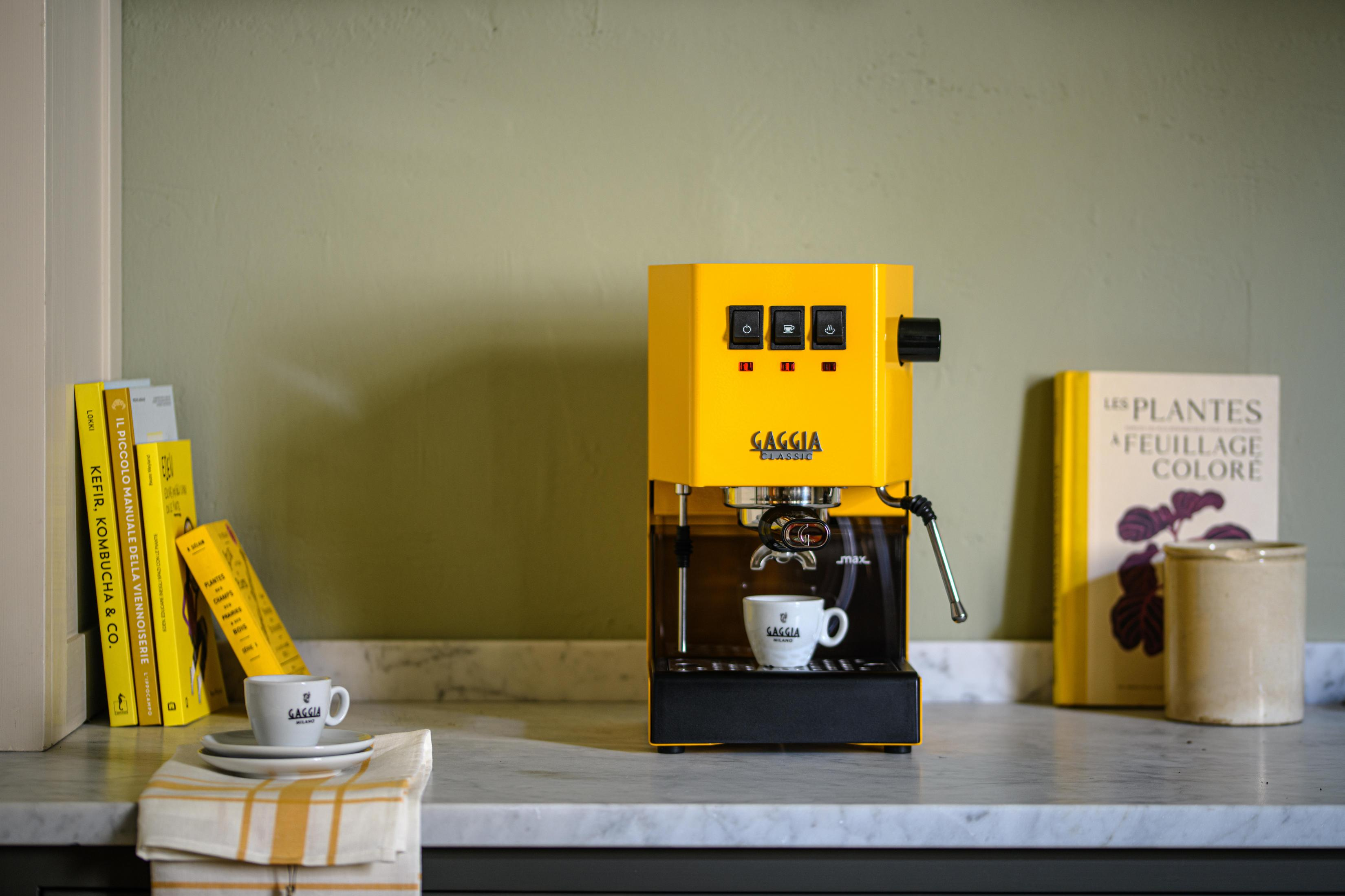 Een gele espressomachine met kopje, boeken en kan op een marmeren aanrecht.