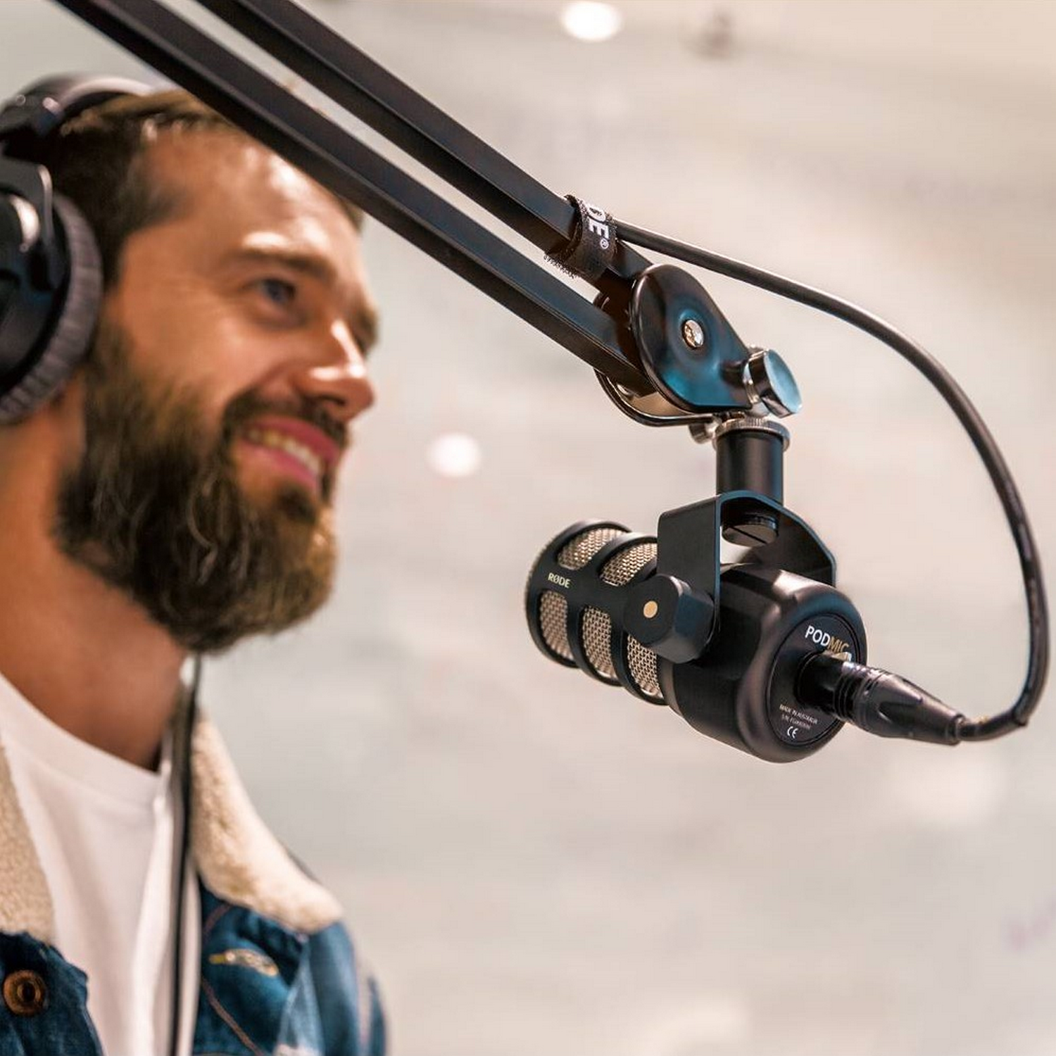 A man with a beard and headphones smiles into a microphone. He wears a denim jacket.