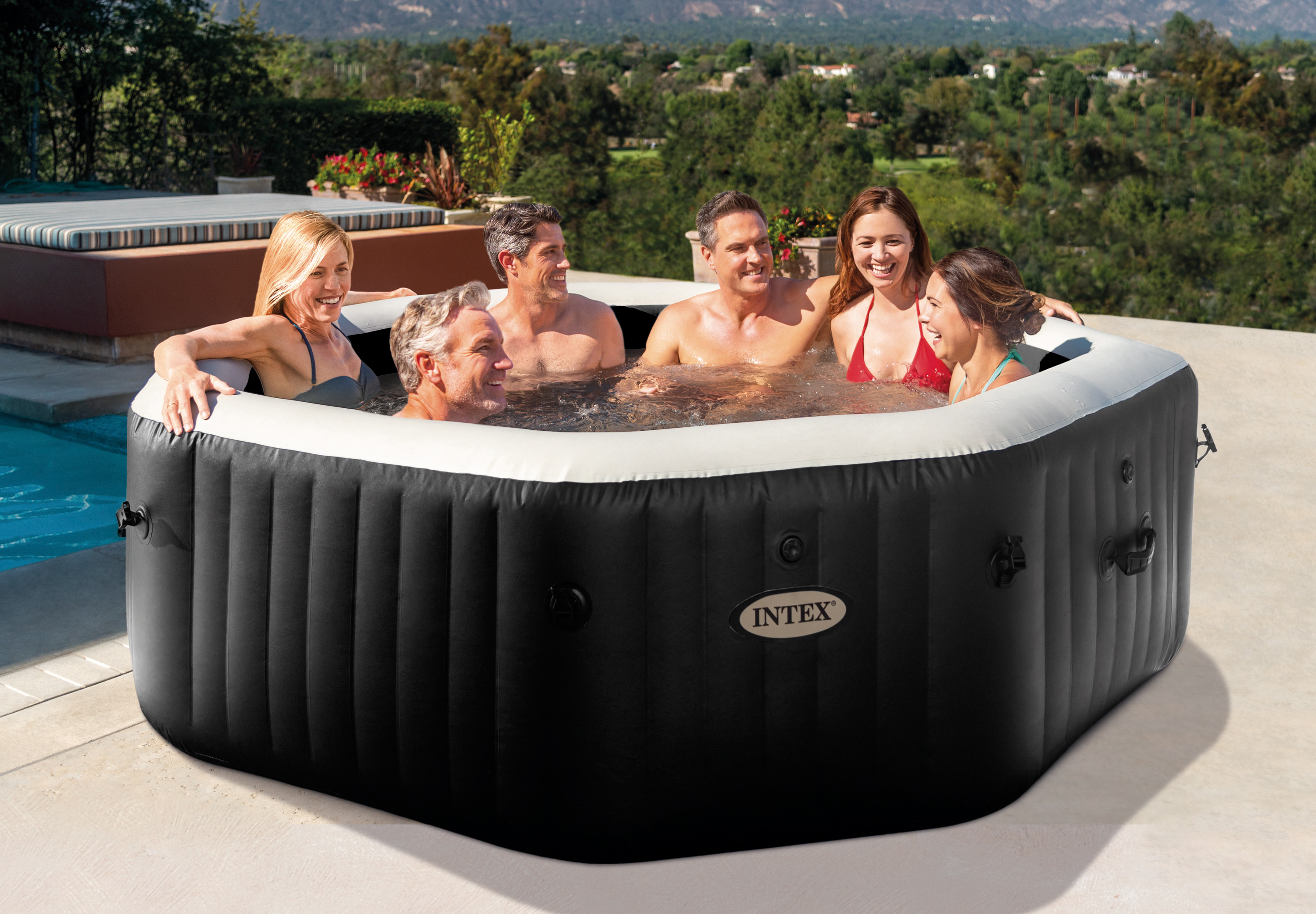 Sechs Personen planschen in einem aufblasbaren Whirlpool im Freien mit Blick auf eine hügelige Landschaft.