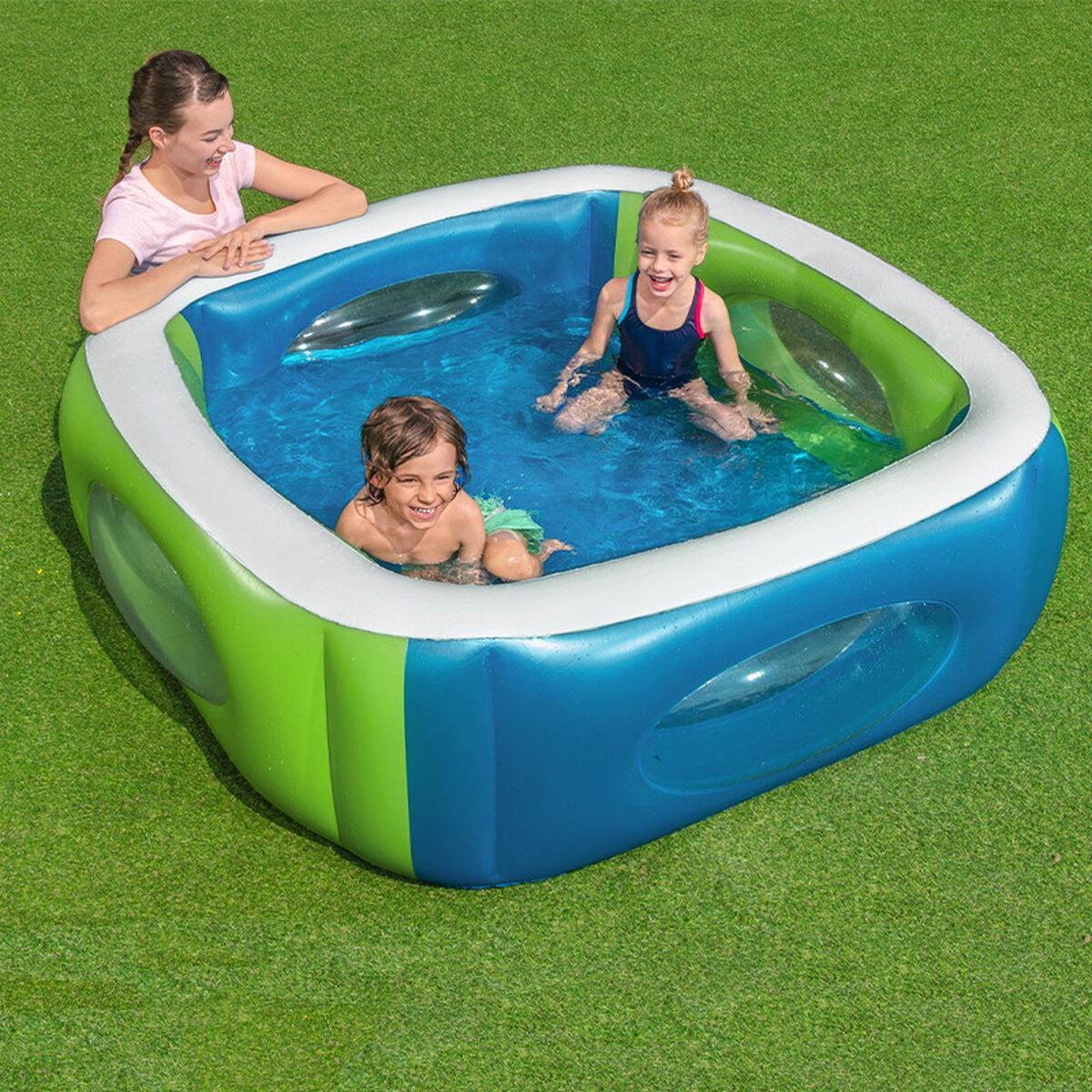 Tres niños, dos dentro y uno inclinado, sonríen y juegan en una piscina inflable azul y verde sobre el césped.