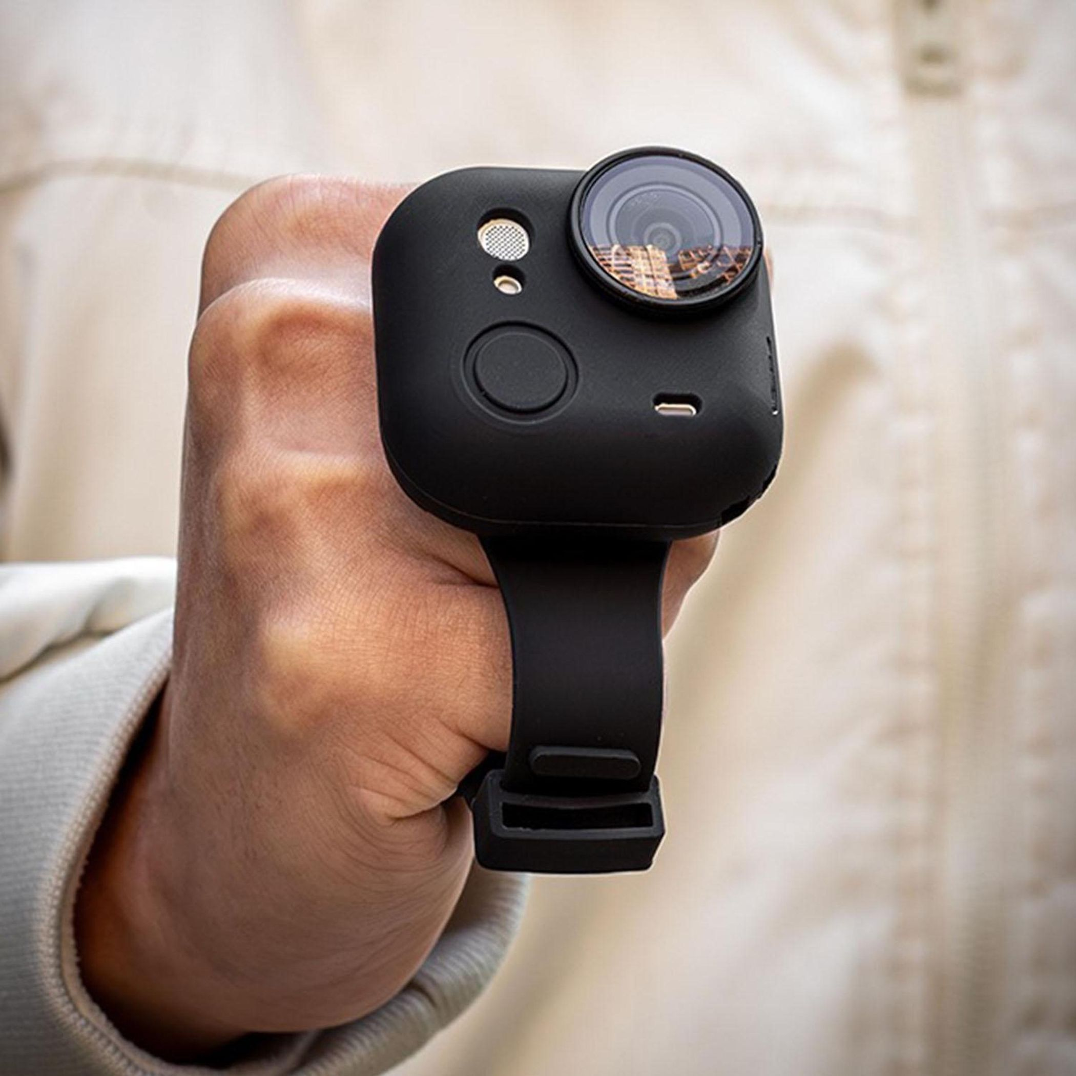 Eine schwarze Kamera mit Objektiv wird in der Hand gehalten, die an einem Armband befestigt ist.
