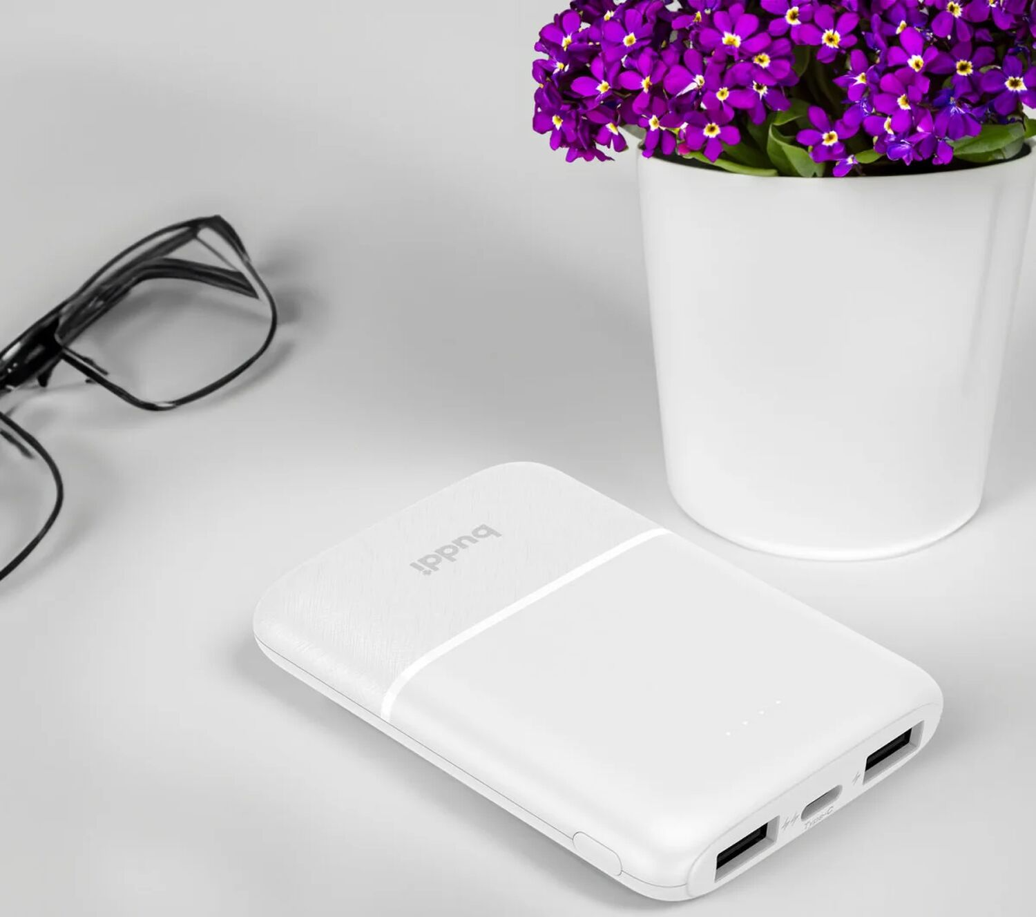Witte powerbank naast bril en paarse bloemen in witte pot.