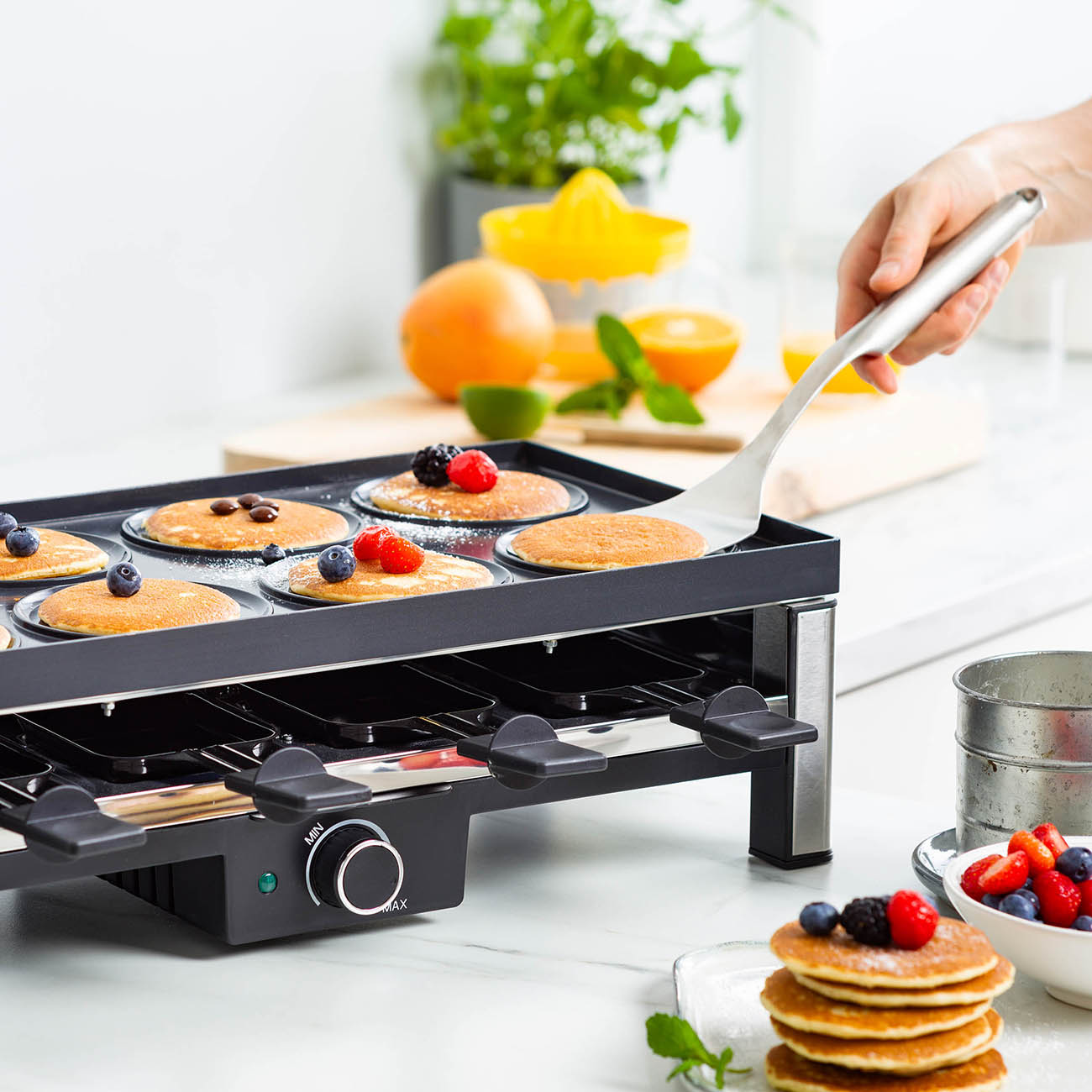 Person macht Pfannkuchen auf einer elektrischen Grillplatte mit Beeren und Obst im Hintergrund.