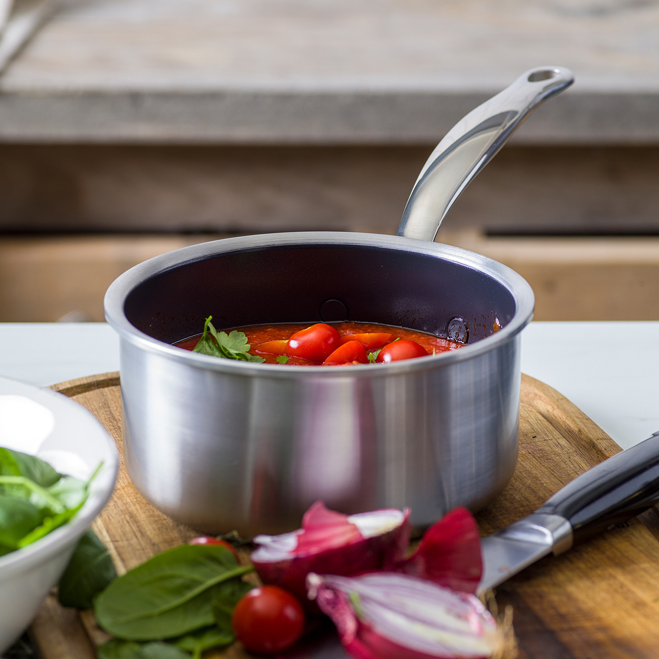 Una olla de salsa de tomate está sobre una tabla de madera junto a cebollas y espinacas.