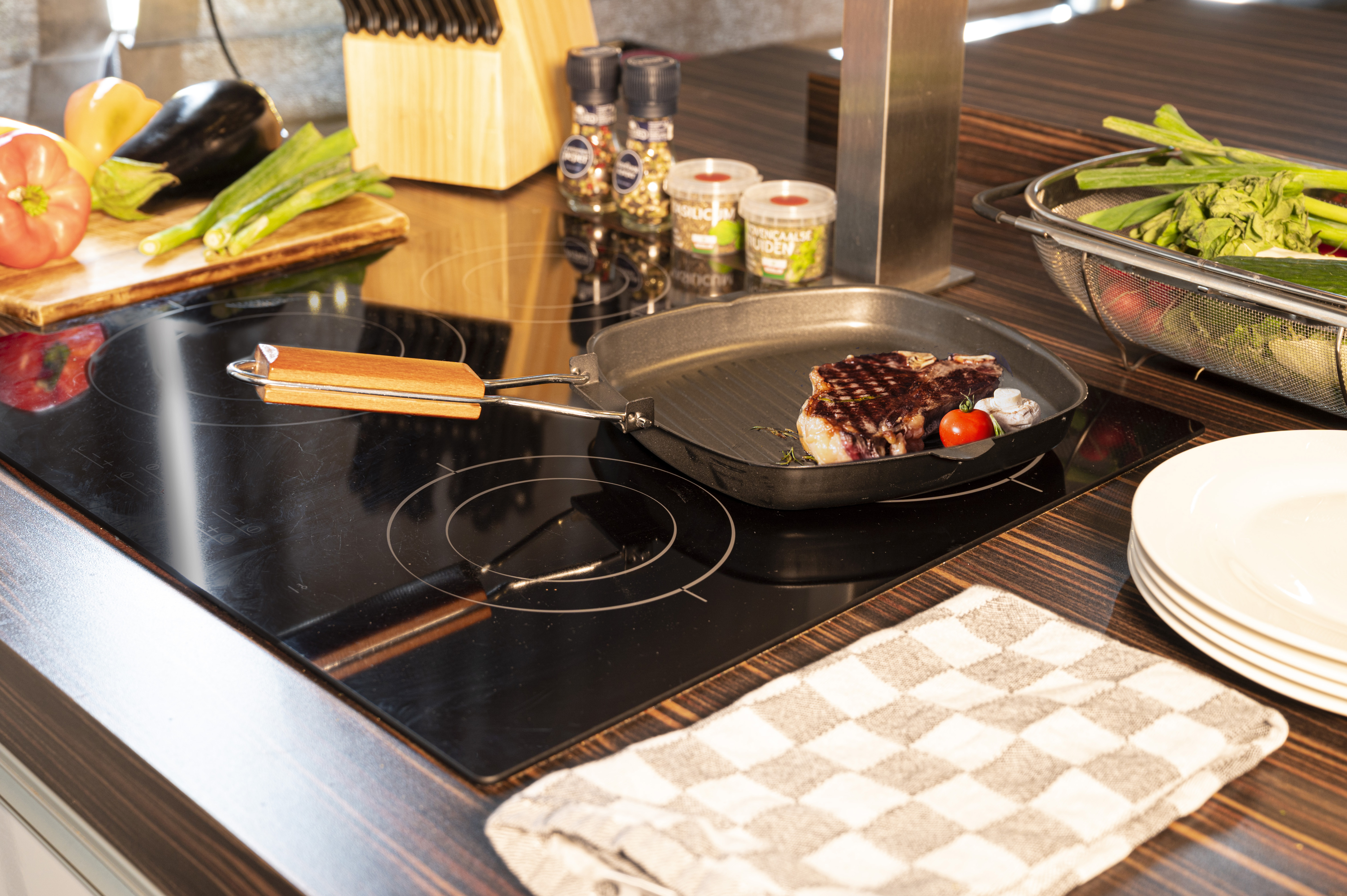 A steak is cooking in a grill pan on a stovetop, with vegetables and spices nearby.