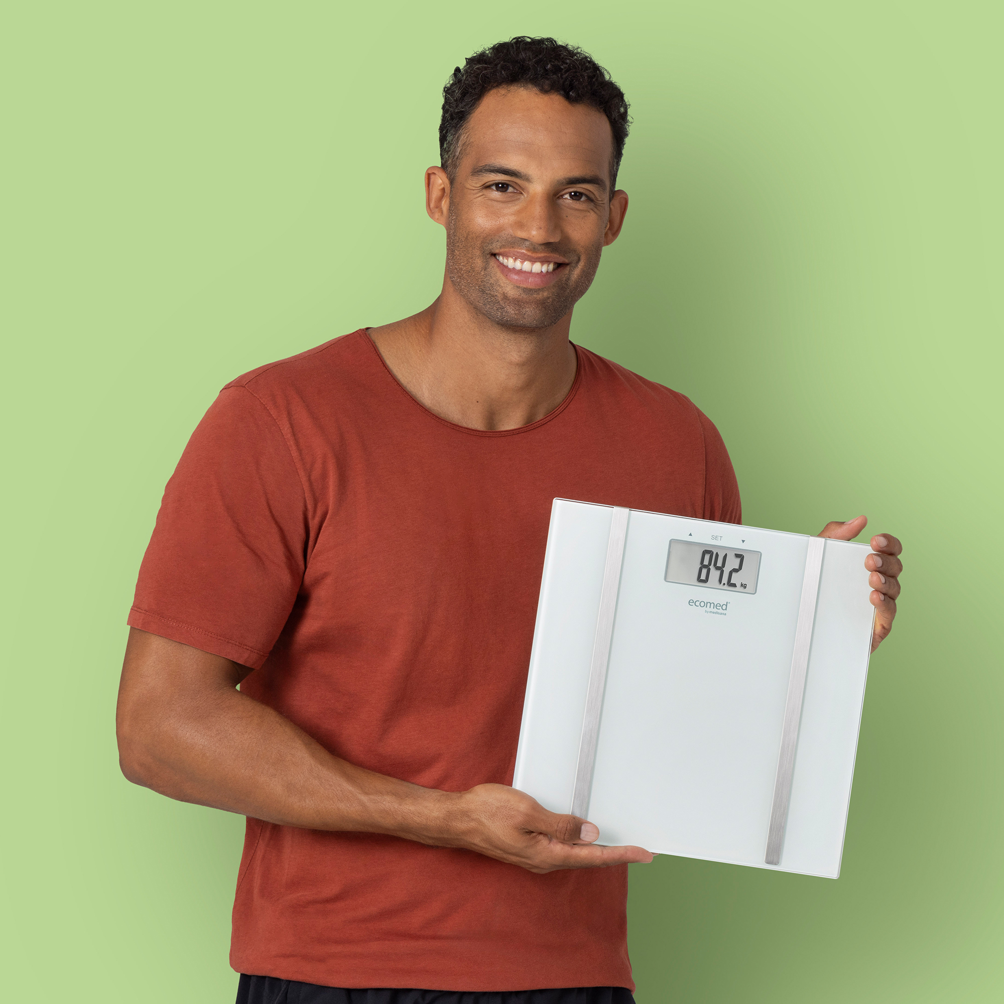 Mann in rotem Hemd hält eine weiße Waage mit Anzeige 84,2 kg.