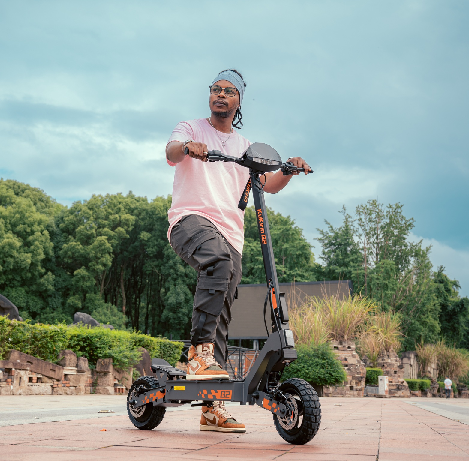 Hombre con camiseta rosa y pantalones cargo monta patinete eléctrico al aire libre con árboles.