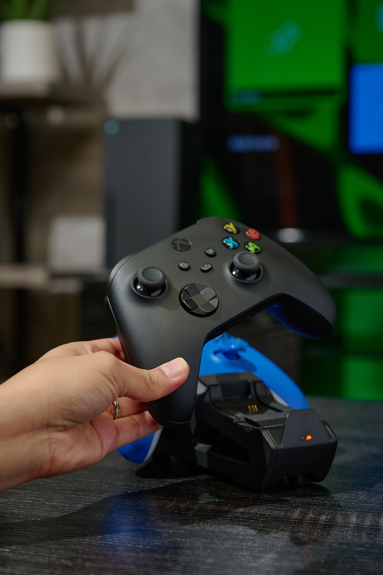 Eine Hand hält einen schwarzen Xbox-Controller über einer blauen Ladestation mit beleuchtetem orangefarbenem Licht.