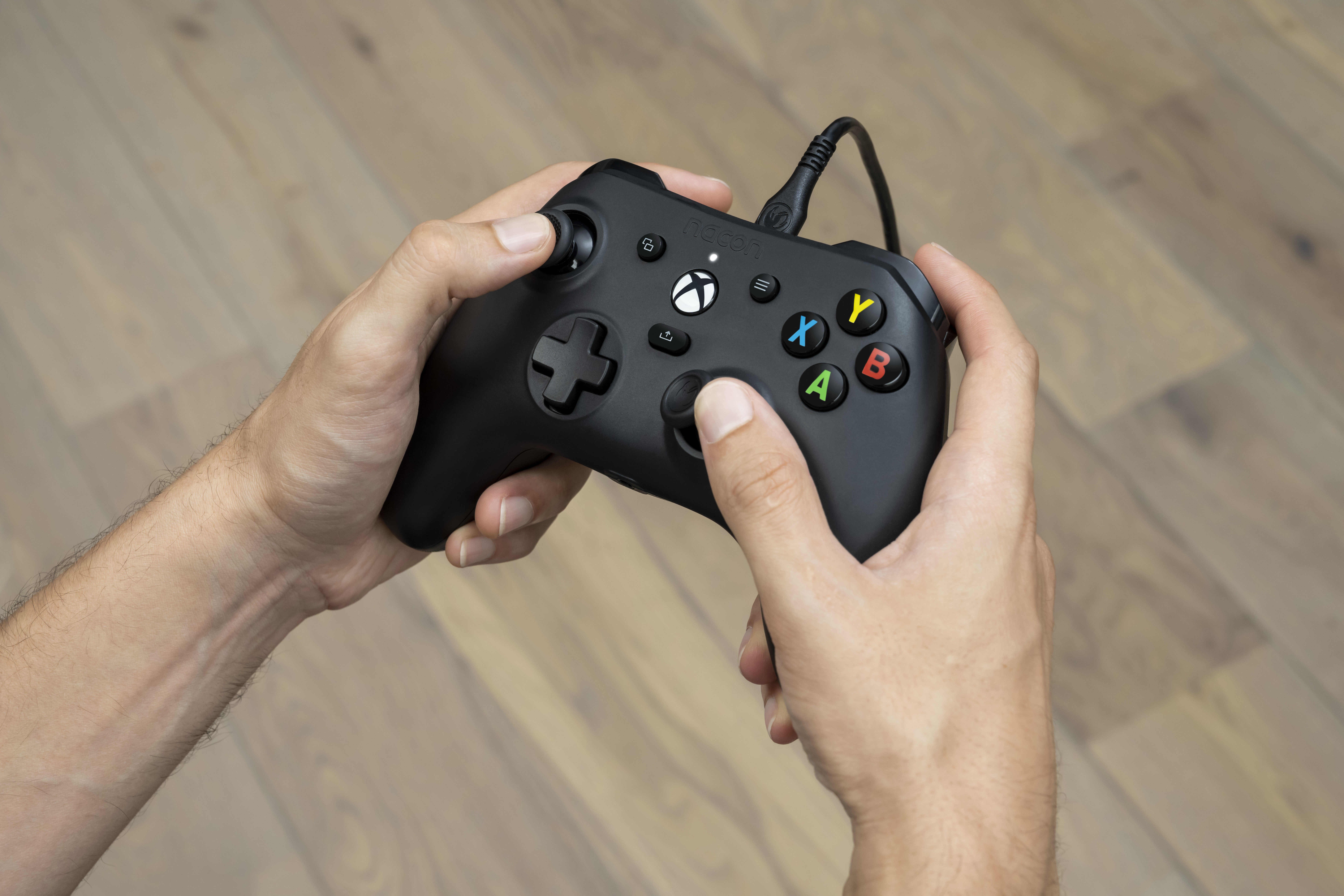Hände halten einen schwarzen Xbox-Controller mit eingestecktem Kabel auf Holzboden.