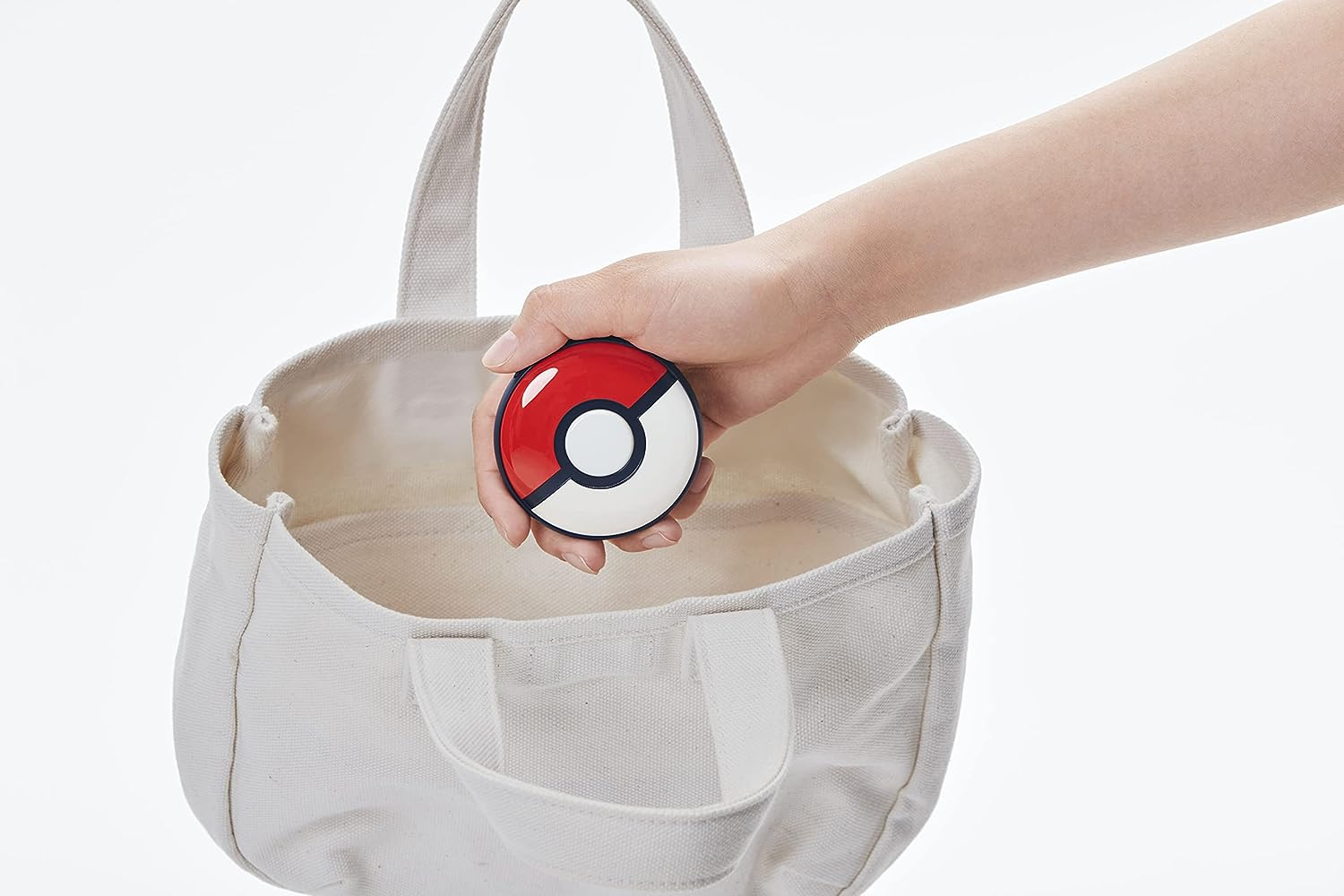 Dłoń trzyma czerwono-białą Pokeballa nad otwartą torbą płócienną.