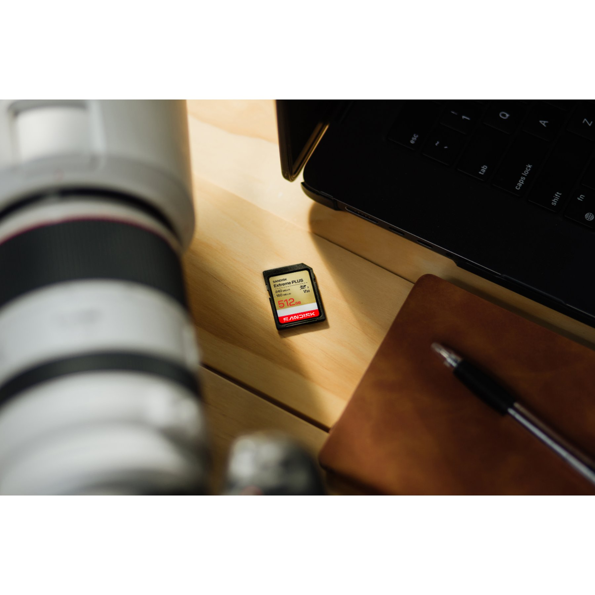 Una scheda SD SanDisk 512GB Extreme Plus poggia su una scrivania in legno accanto all'obiettivo di una fotocamera e un laptop.