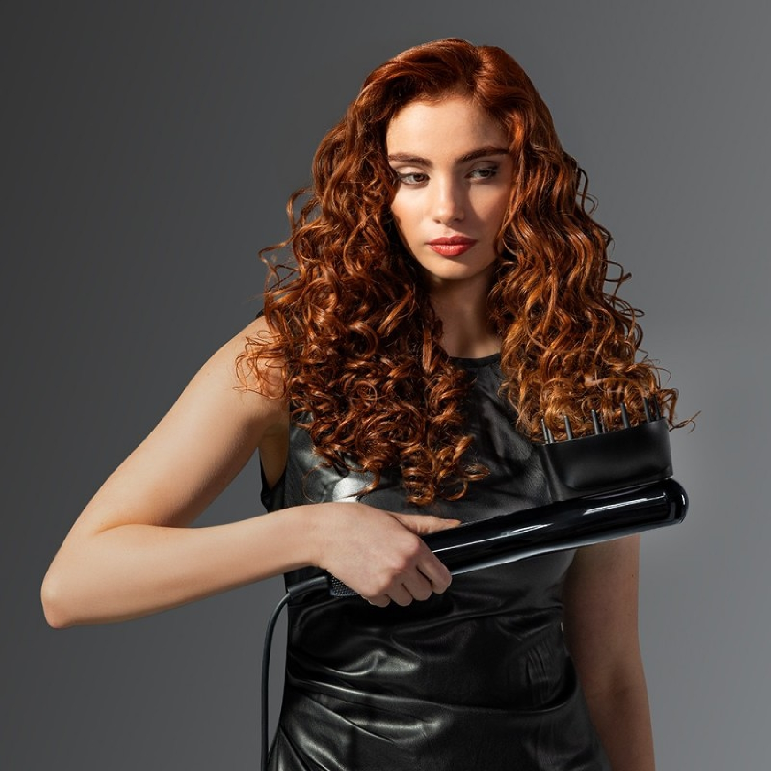 Donna con capelli rossi ricci in abito nero tiene un ferro arricciacapelli.