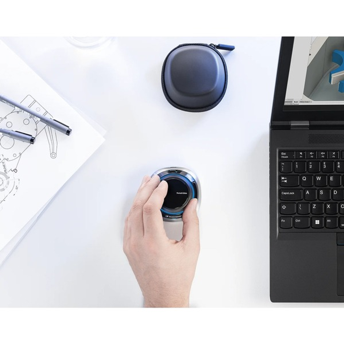 A hand operates a 3D mouse next to a laptop and technical drawings.