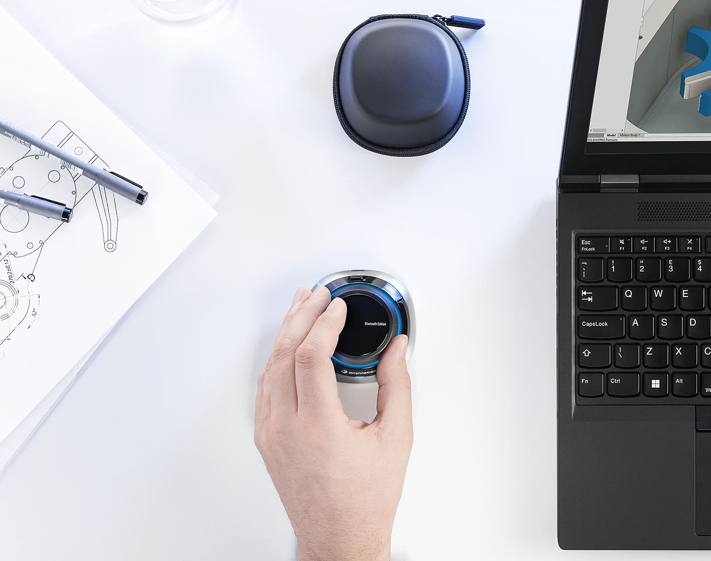 A hand operates a 3D mouse next to a laptop, blueprints, and a case.