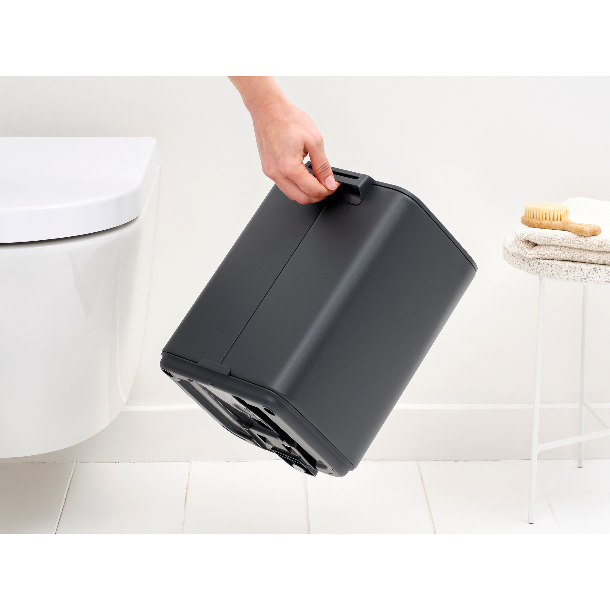 Een hand houdt een donkere prullenbak naast een wit toilet vast.