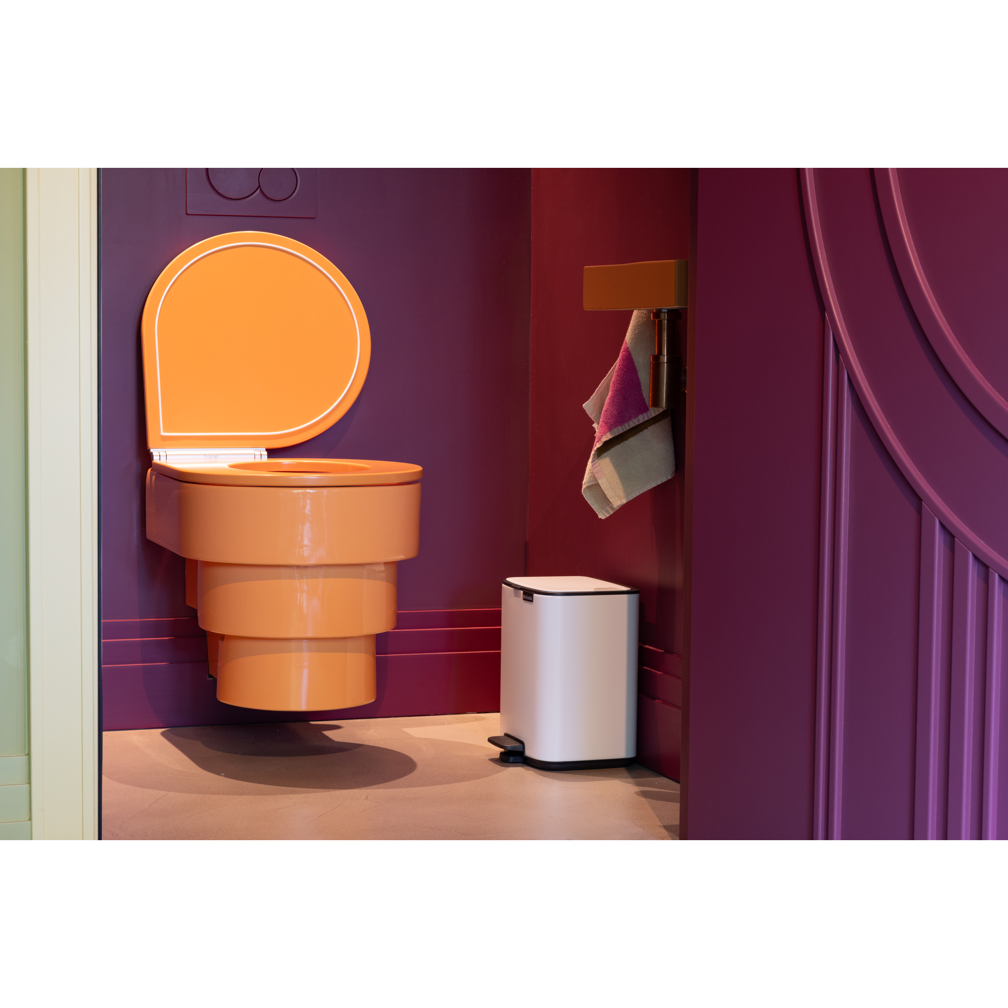 Een oranje toilet met de deksel omhoog, naast een witte prullenbak.