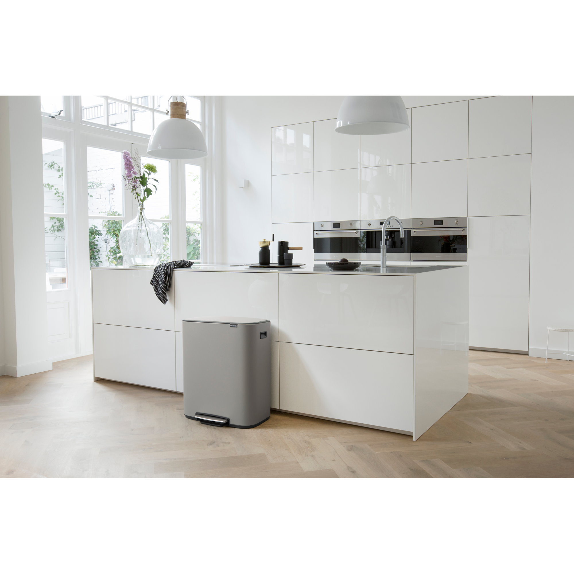 Een moderne witte keuken met eiland, prullenbak en bloemen.
