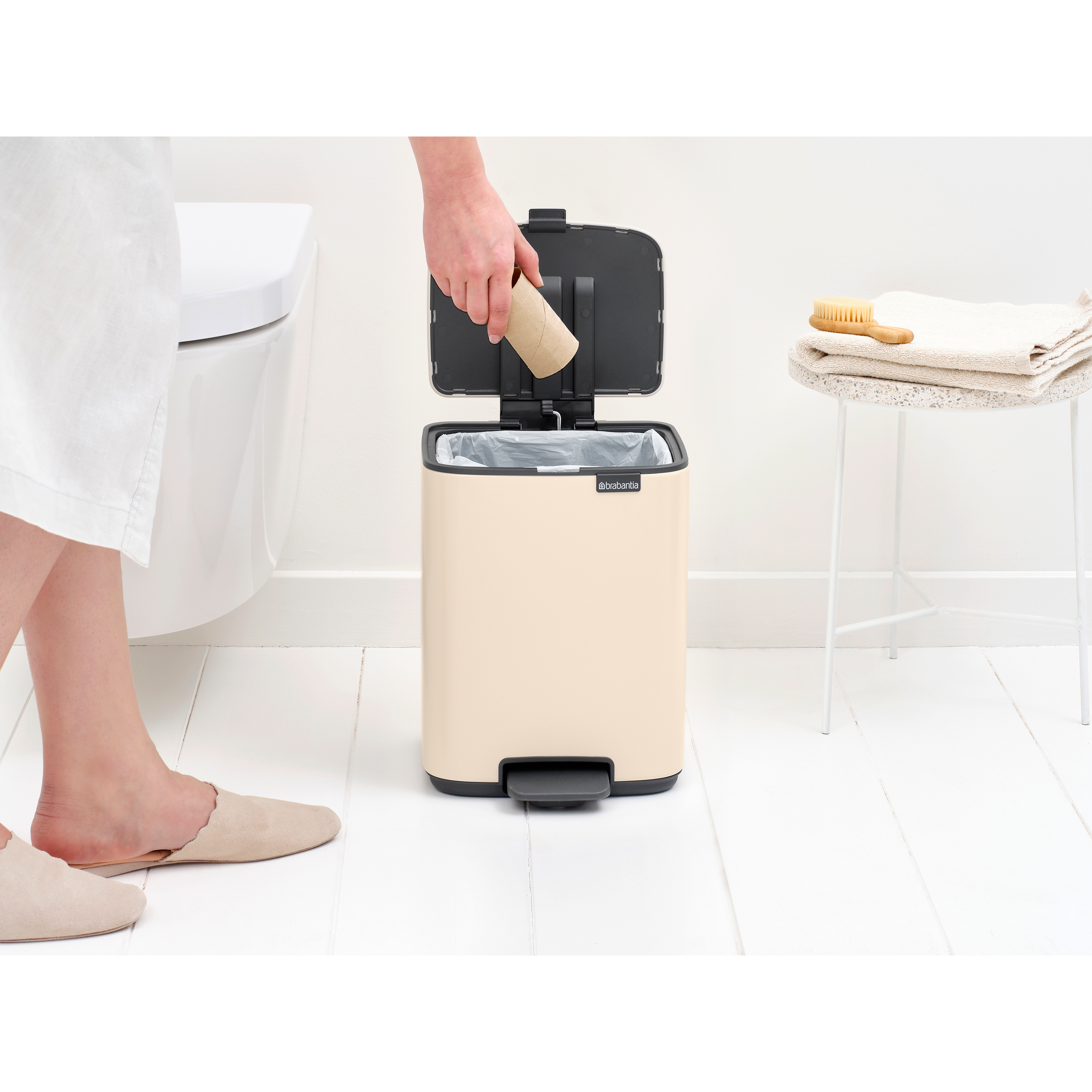 Een hand gooit een kartonnen rol in een crème kleurige prullenbak naast een toilet.