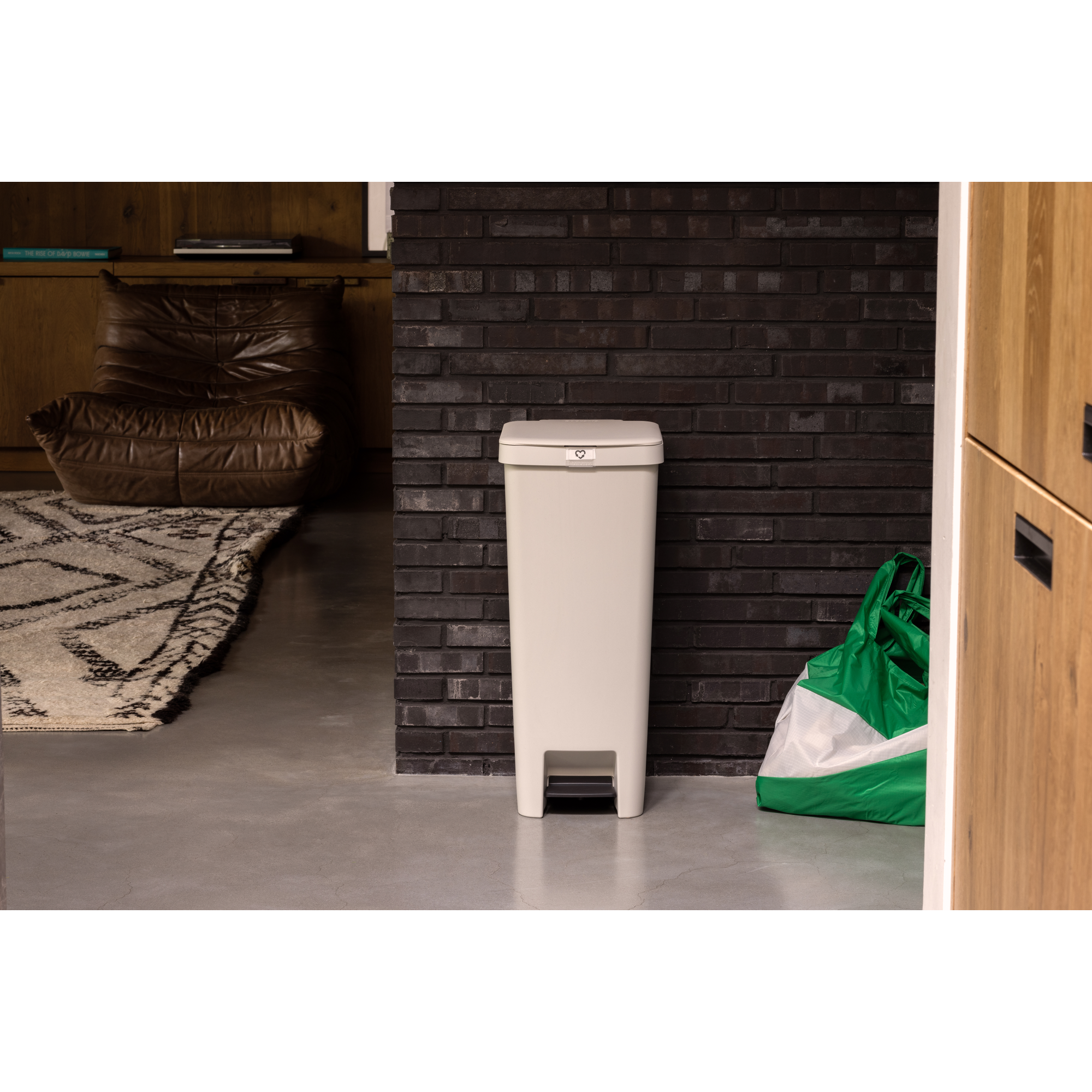 A light-colored trash can with a pedal stands in front of a dark brick wall.