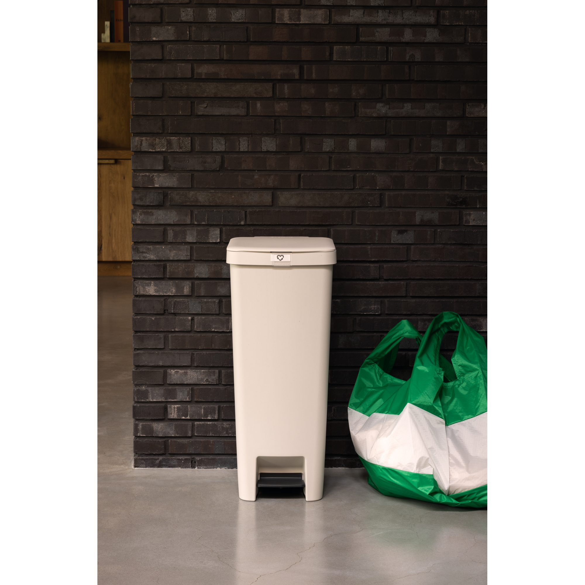 A light-colored pedal trash can next to a green and white bag against a brick wall.