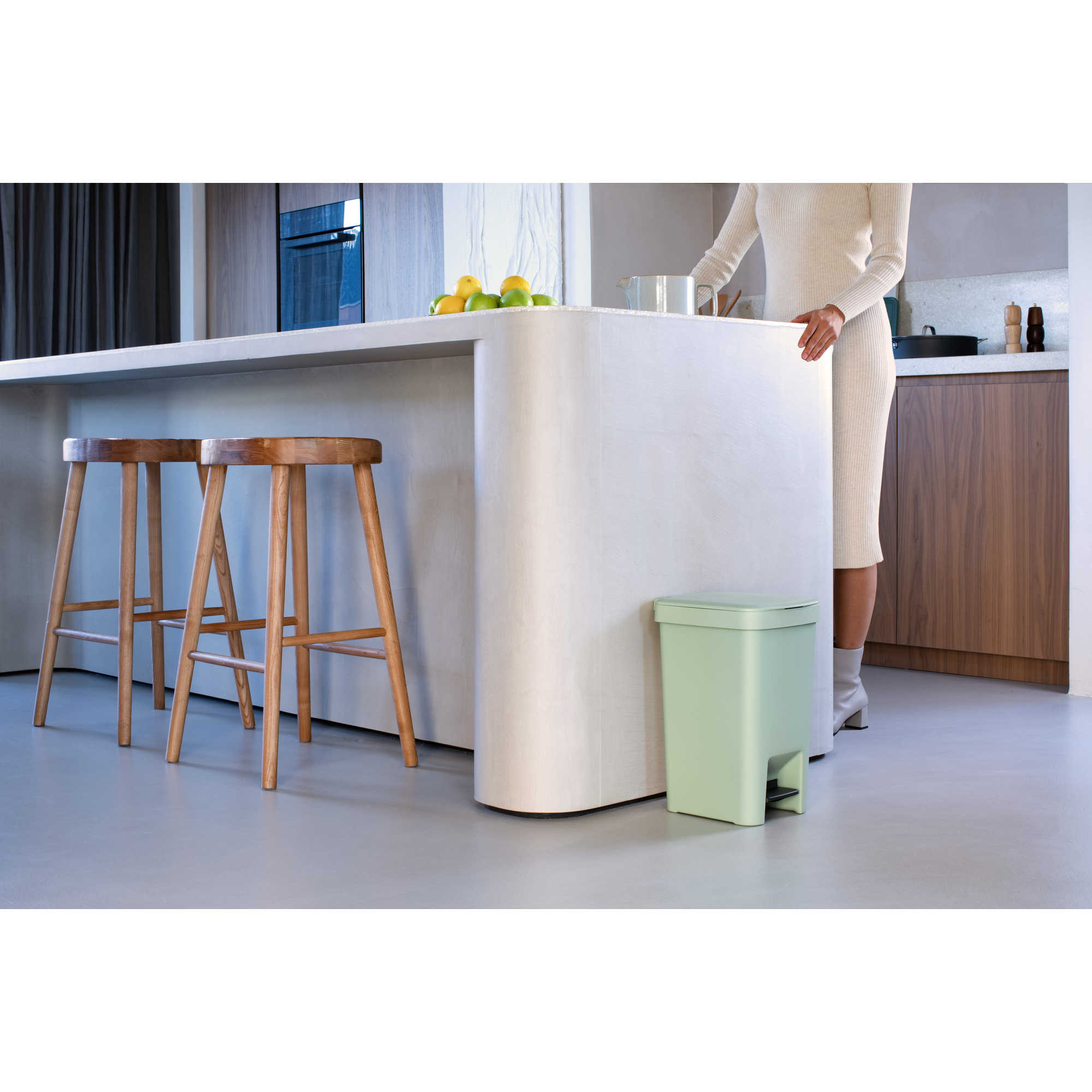 Poubelle à pédale verte près d'un îlot de cuisine avec tabourets et fruits.
