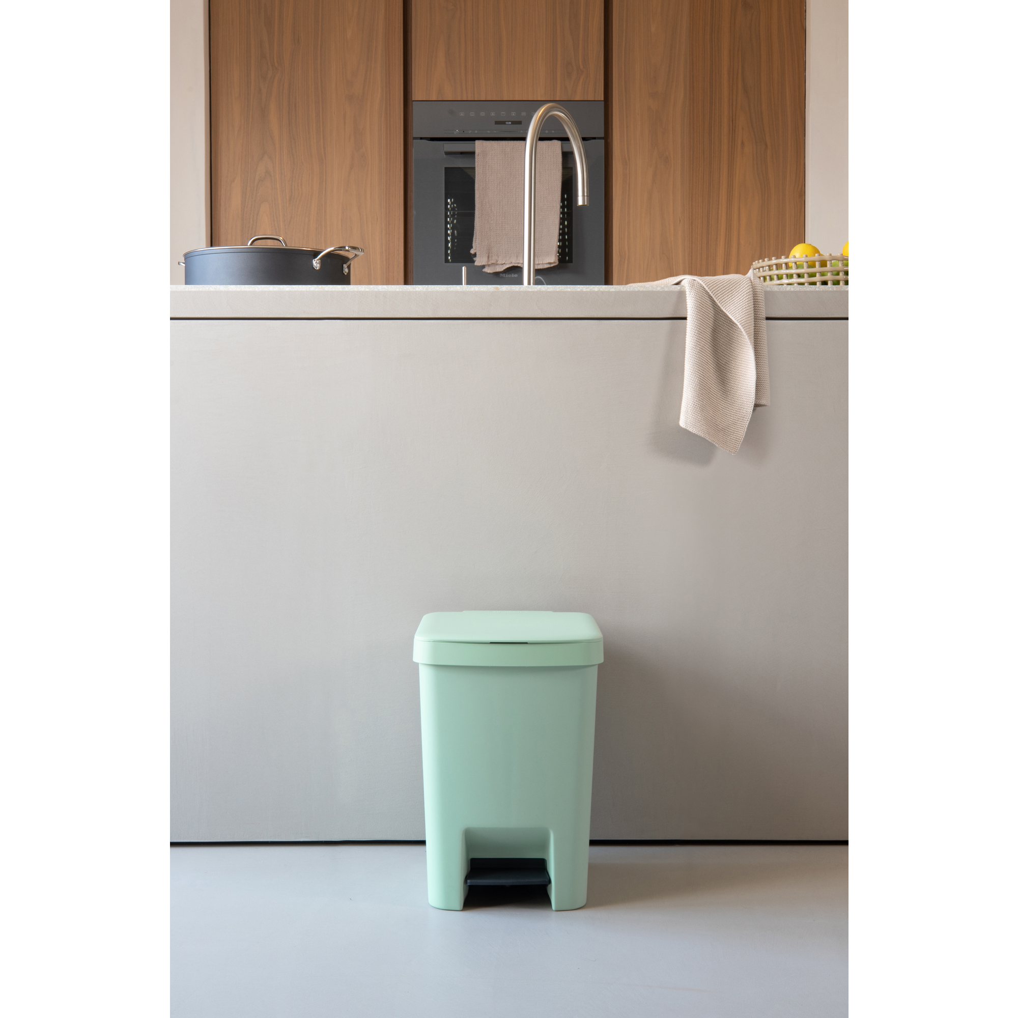 Een mintgroene pedaalemmer staat voor een keukenblad met houten kasten.