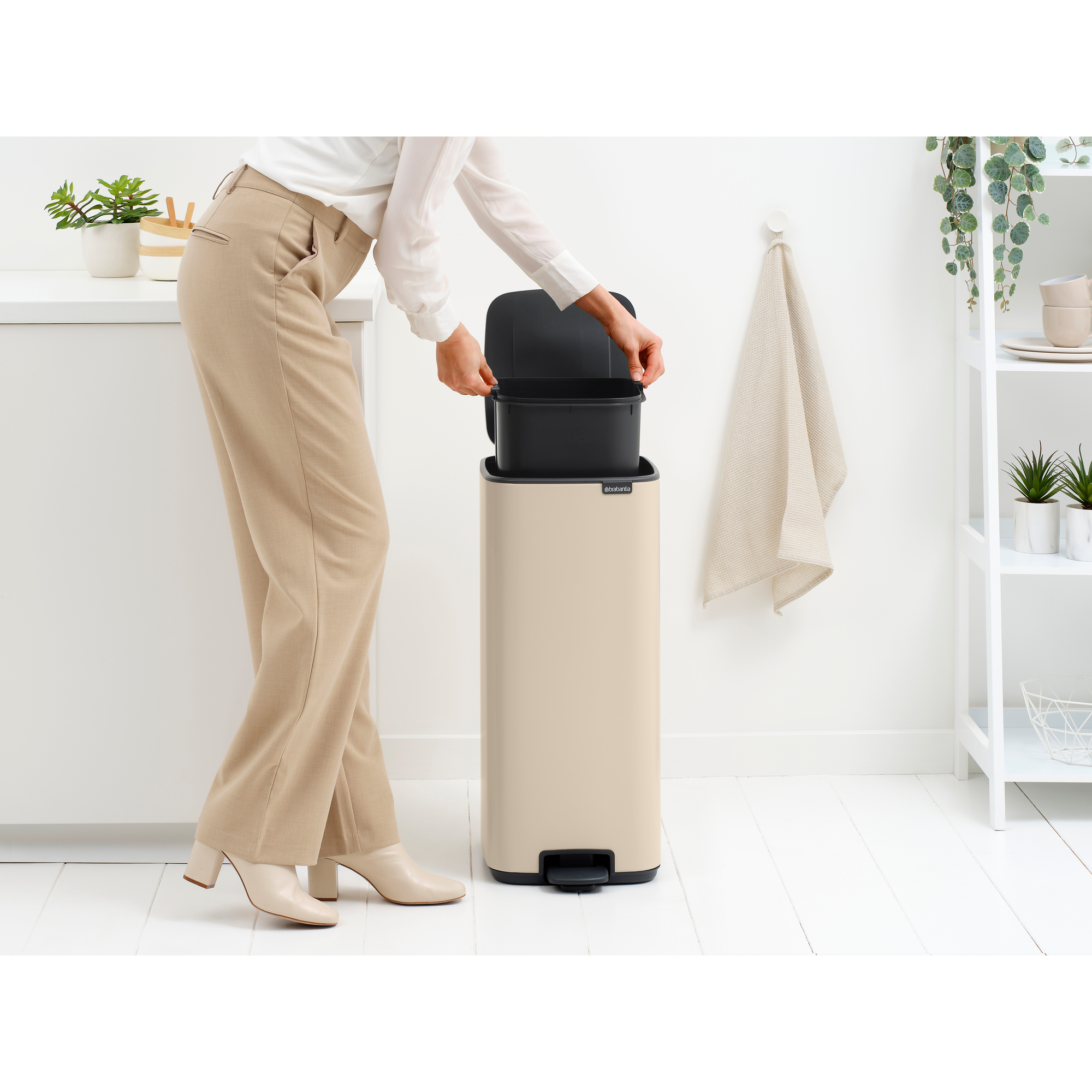 Een vrouw in beige broek en witte blouse opent een zandkleurige prullenbak met zwart inzetstuk.