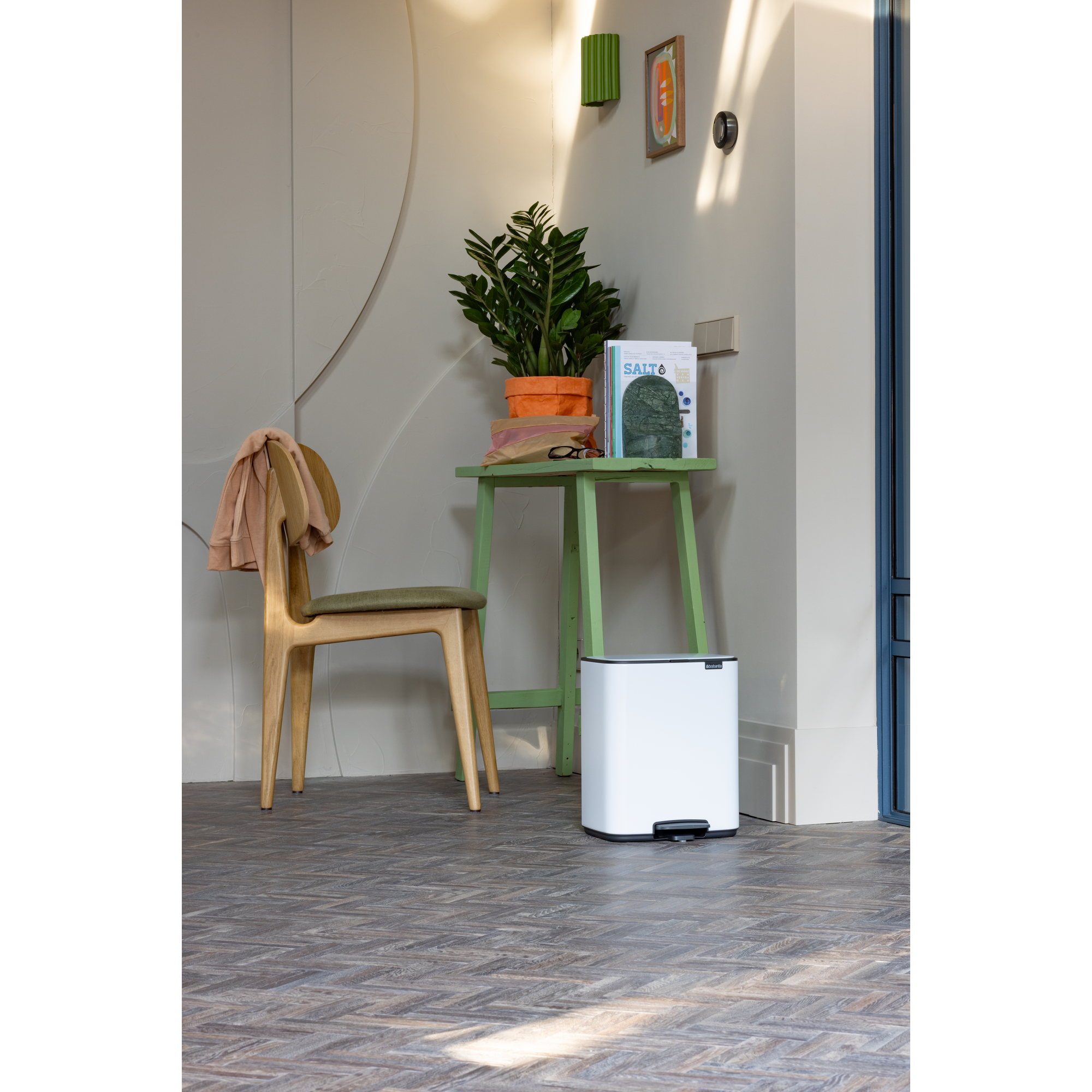 Een witte pedaalemmer staat naast een groen bijzettafeltje met plant, boeken en een bril.