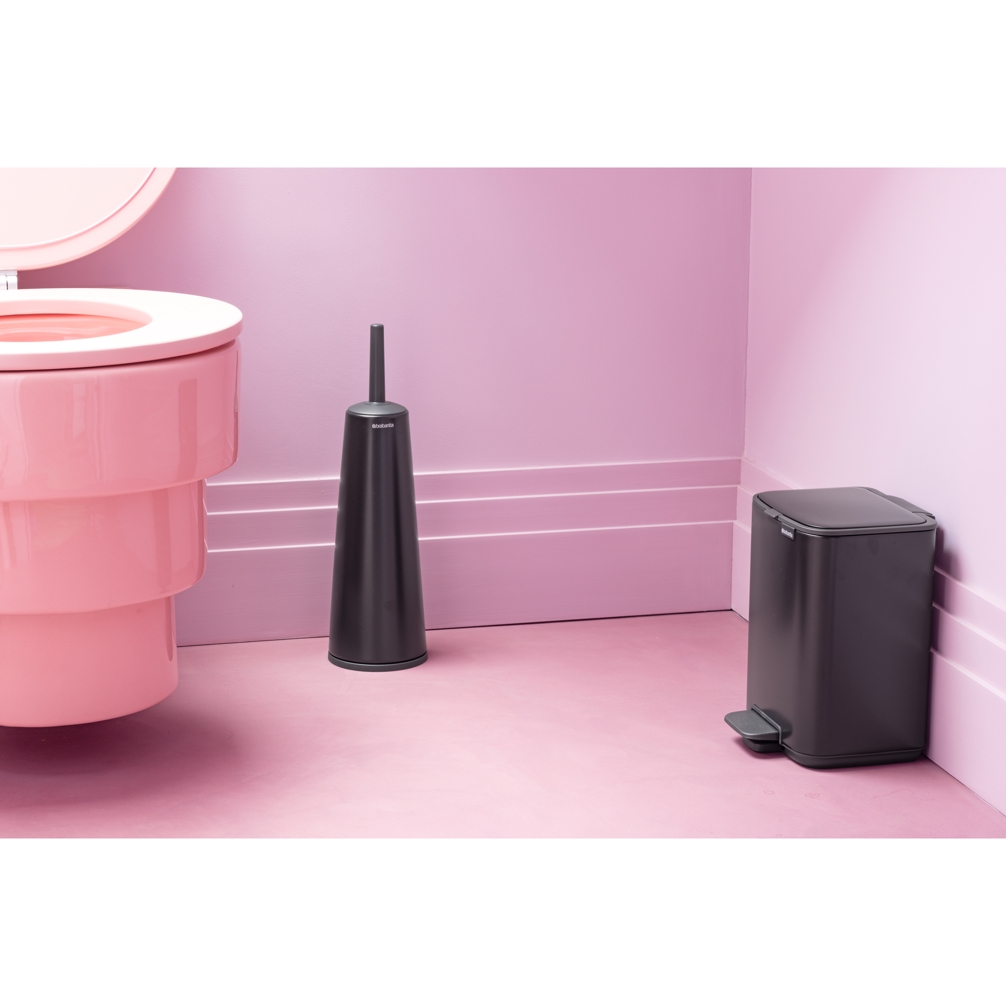 Een roze toilet, een zwarte toiletborstel en een zwarte prullenbak op roze vloer.