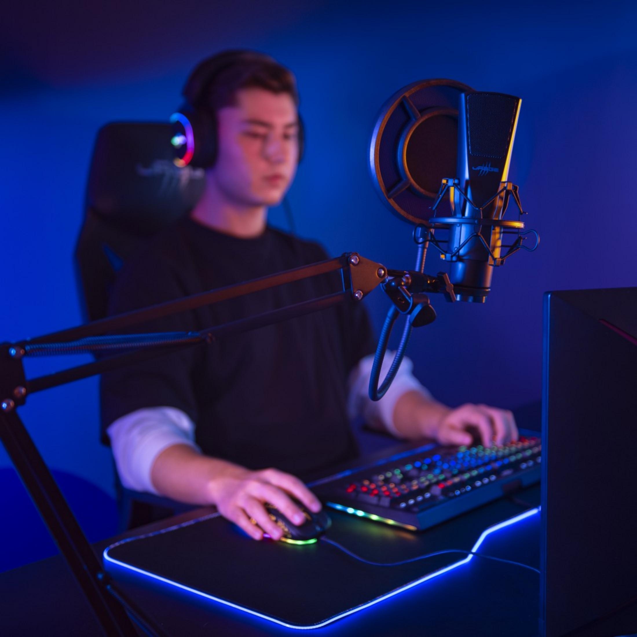 Ein Mann mit Kopfhörern sitzt vor einem beleuchteten Mikrofon und spielt am Computer.